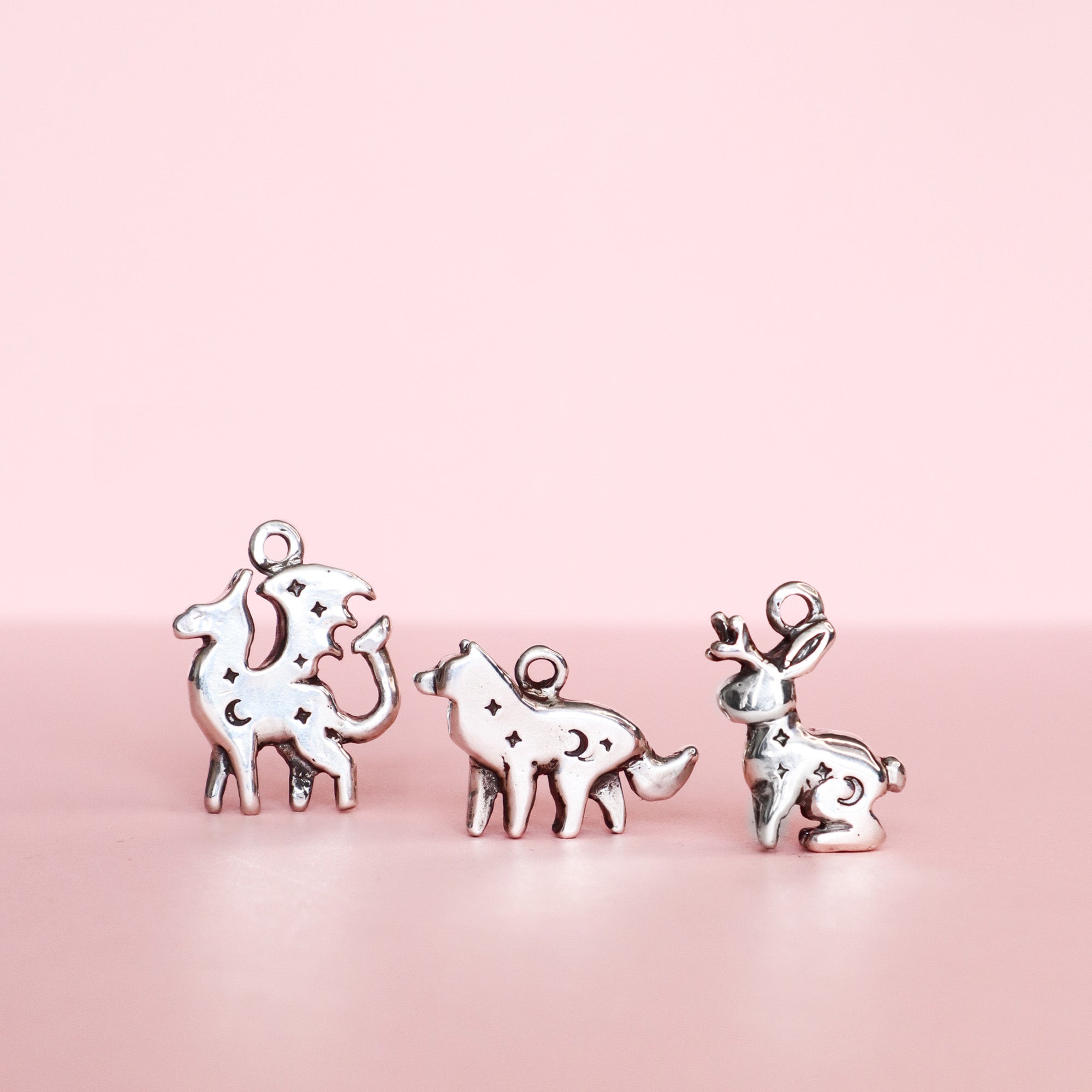 The Silver Lunar Wolf Pendant by Papio Press, a sterling silver charm with delicate star-shaped cutouts, is displayed on a pink surface and background alongside animal-themed pendants.