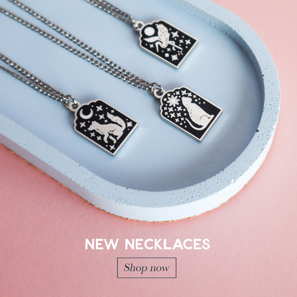 A photograph of 3 silver necklaces on a blue tray. One necklace features a wolf, one a cat and the last features a crane.