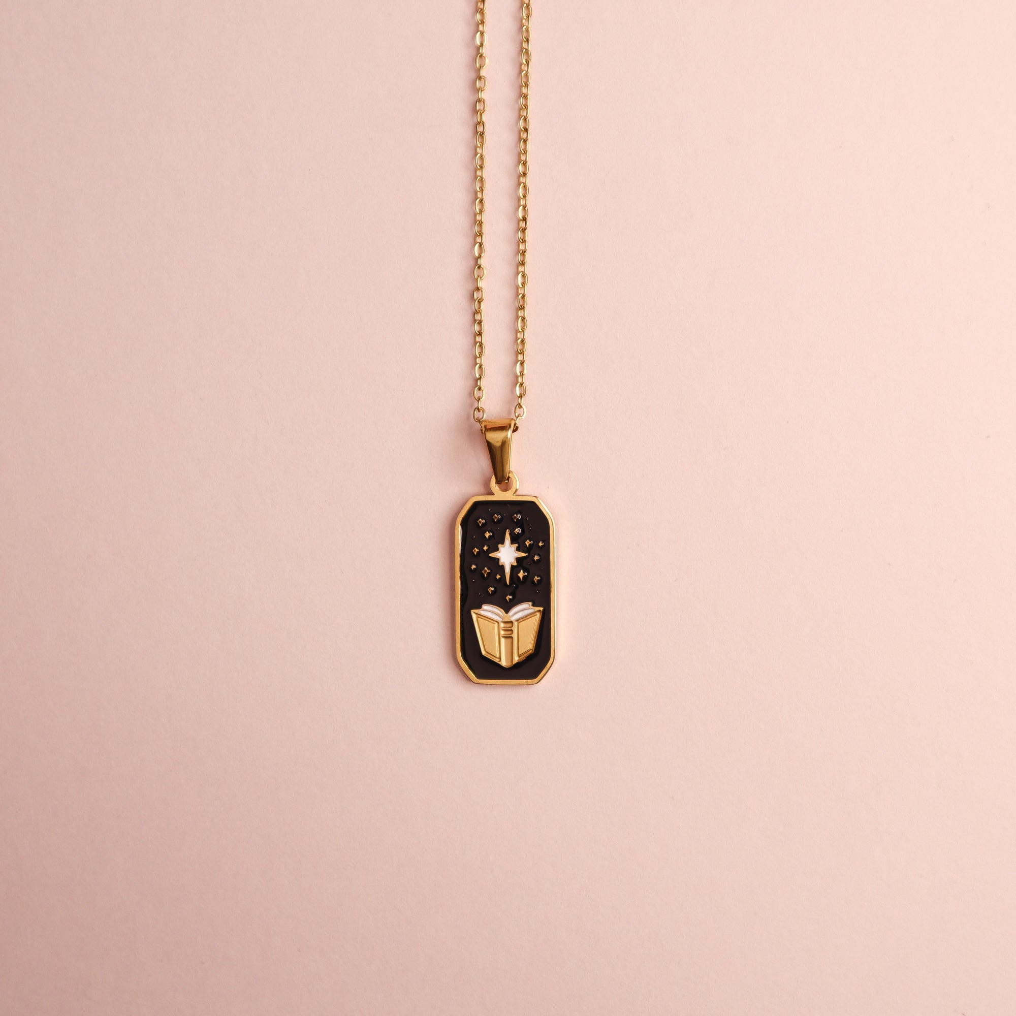The Sorcerer Pendant Enamel Necklace by Papio Press features a gold rectangular pendant with an open book and star design on a black background—an enchanting piece of fantasy jewelry displayed on a pale pink surface.