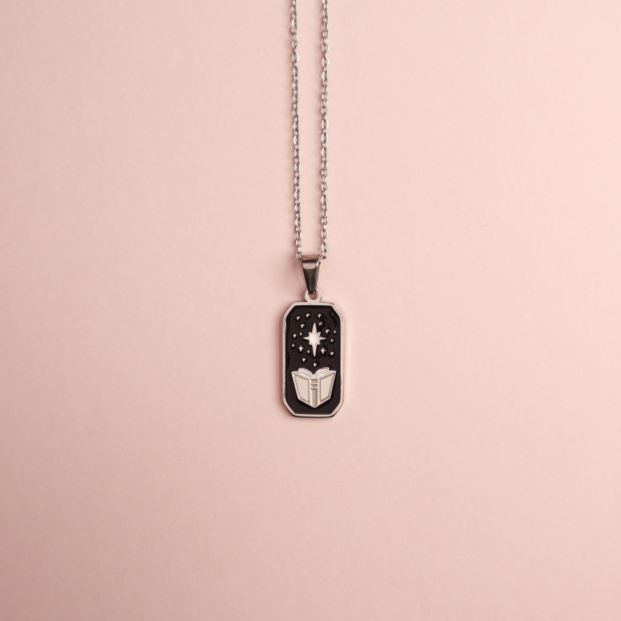 The Sorcerer Pendant Enamel Necklace by Papio Press features a rectangular silver spell book pendant with an open book and white star on black, displayed on light pink—ideal for fantasy jewelry fans.