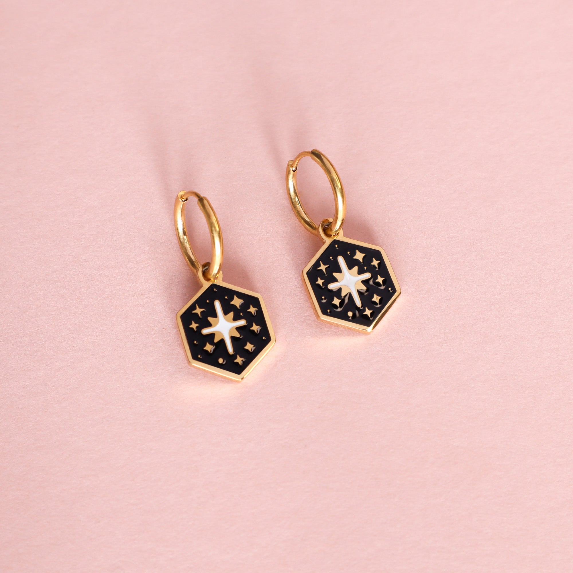 Papio Press Mix and Match Mini Hexagon Earrings: gold plated huggie hoops with black enamel hexagonal charms, white sun design, and gold stars, displayed on a light pink background.
