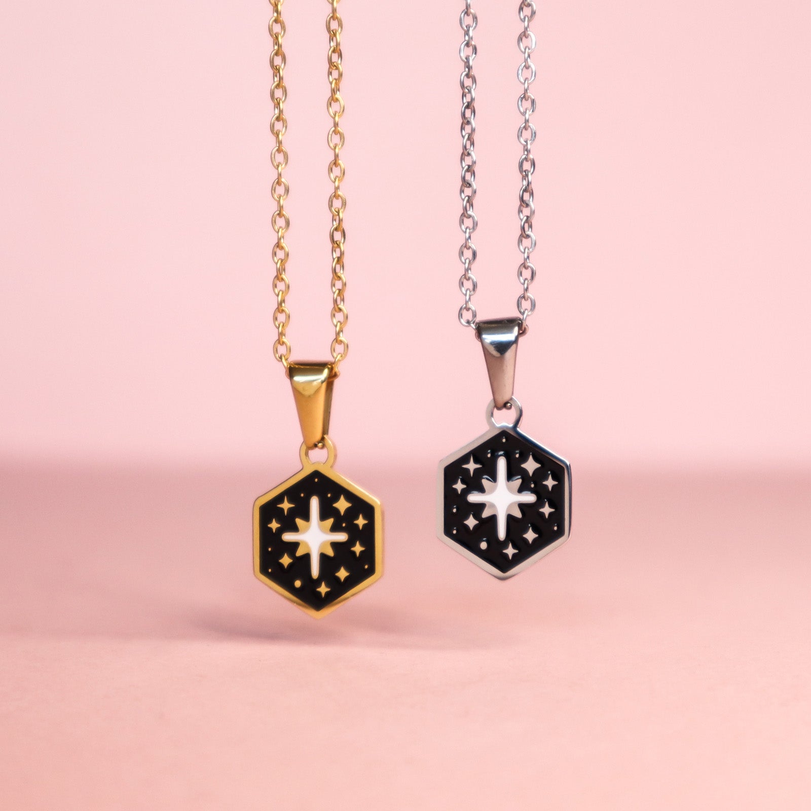 Two Mini Hexagon Star Pendants by Papio Press, with black enamel centers, hang from gold and silver stainless steel chains and feature intricate detailing. Necklaces are displayed against a pink background for a modern look.
