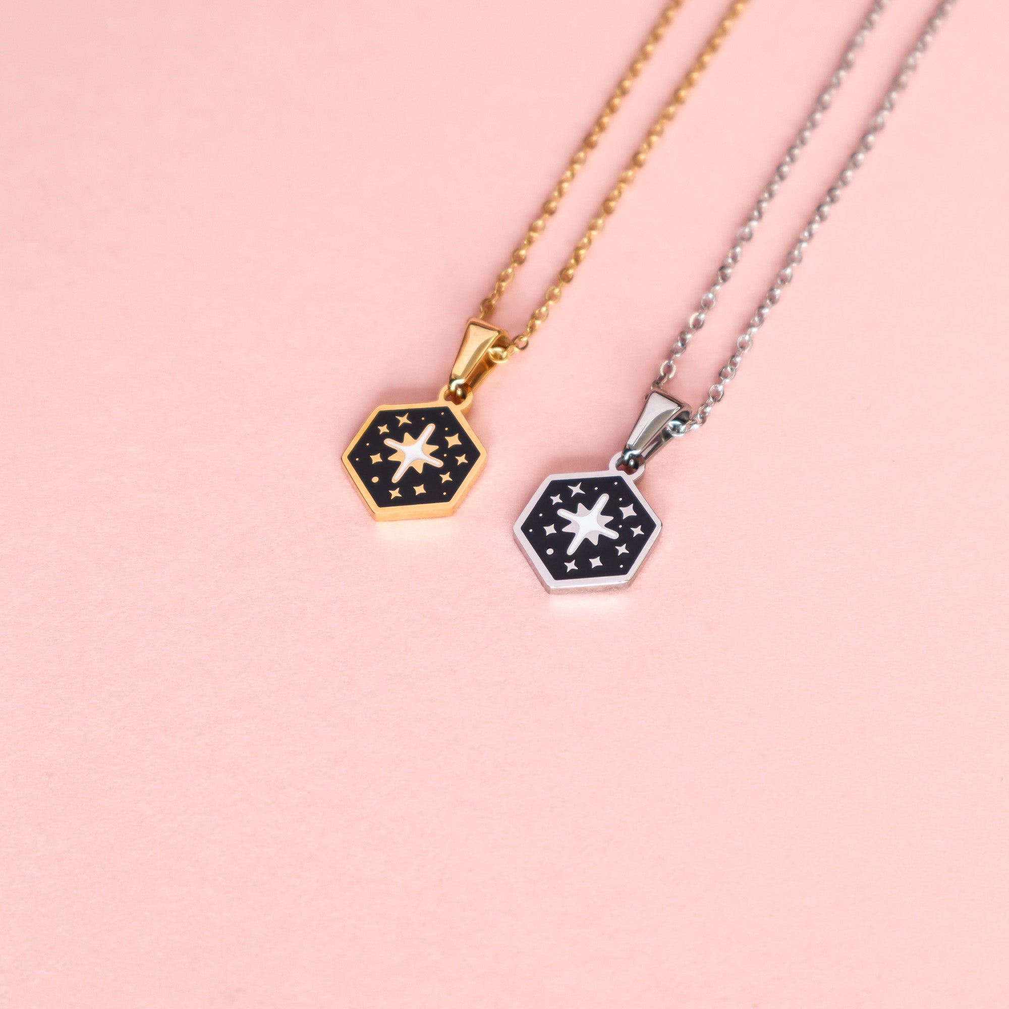 Two Mini Hexagon Star Pendants by Papio Press—one gold, one silver—showcase a white insect and stars on black enamel. Crafted from stainless steel, these necklaces shine on a pink background as a striking modern duo.