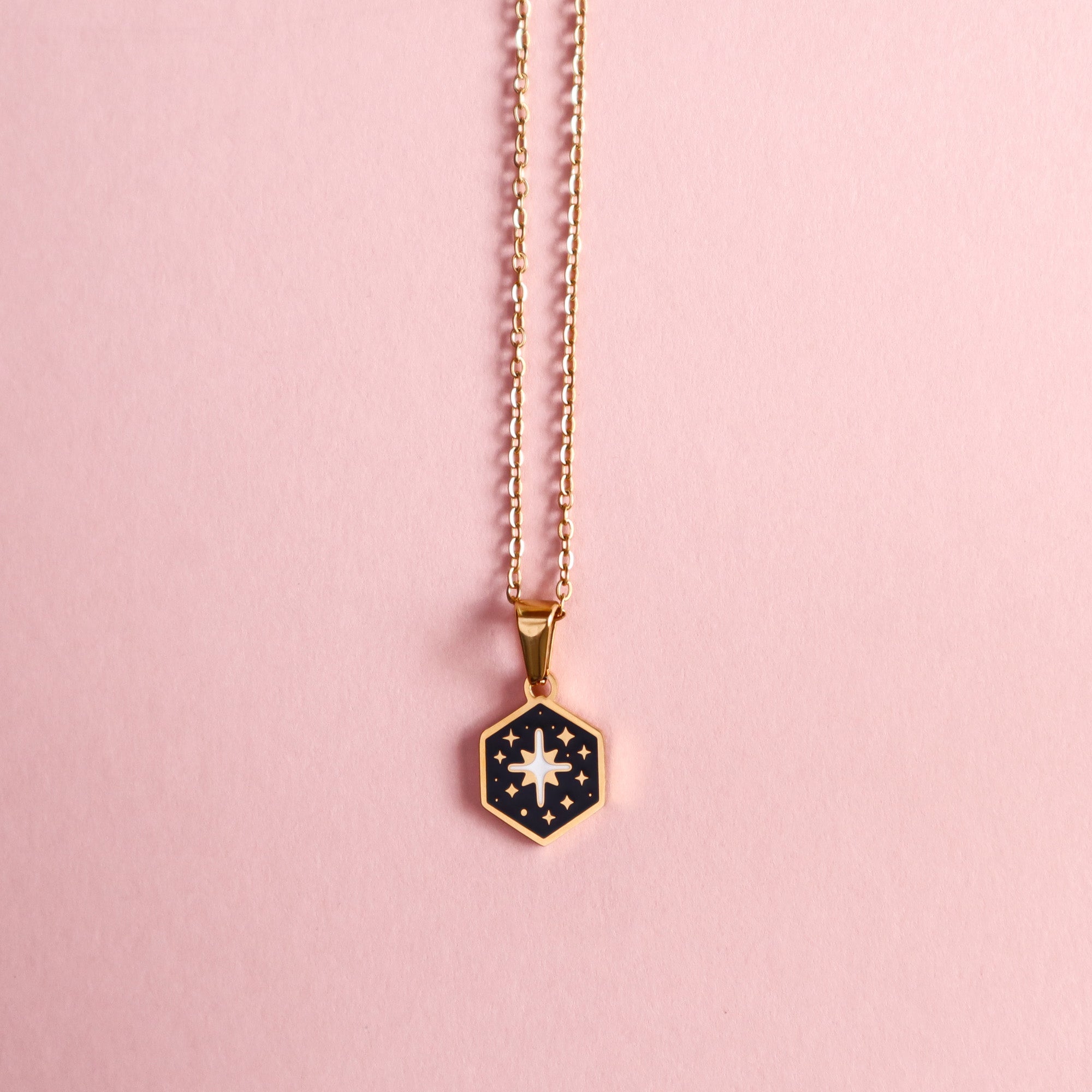 The Mini Hexagon Star Pendant by Papio Press is a gold enamel necklace with a black background, gold stars, and a white starburst design. It’s shown on a pink surface.