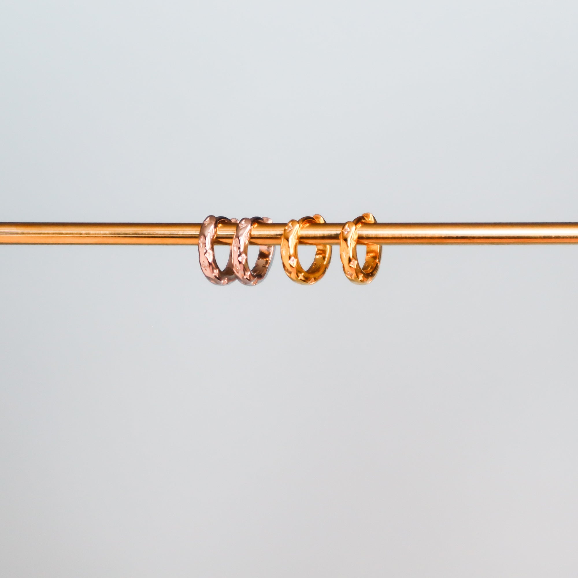 Three Papio Press Star Huggie Hoop Earrings—two in gold and one in rose gold—are displayed side by side on a thin gold rod against a plain light grey background.