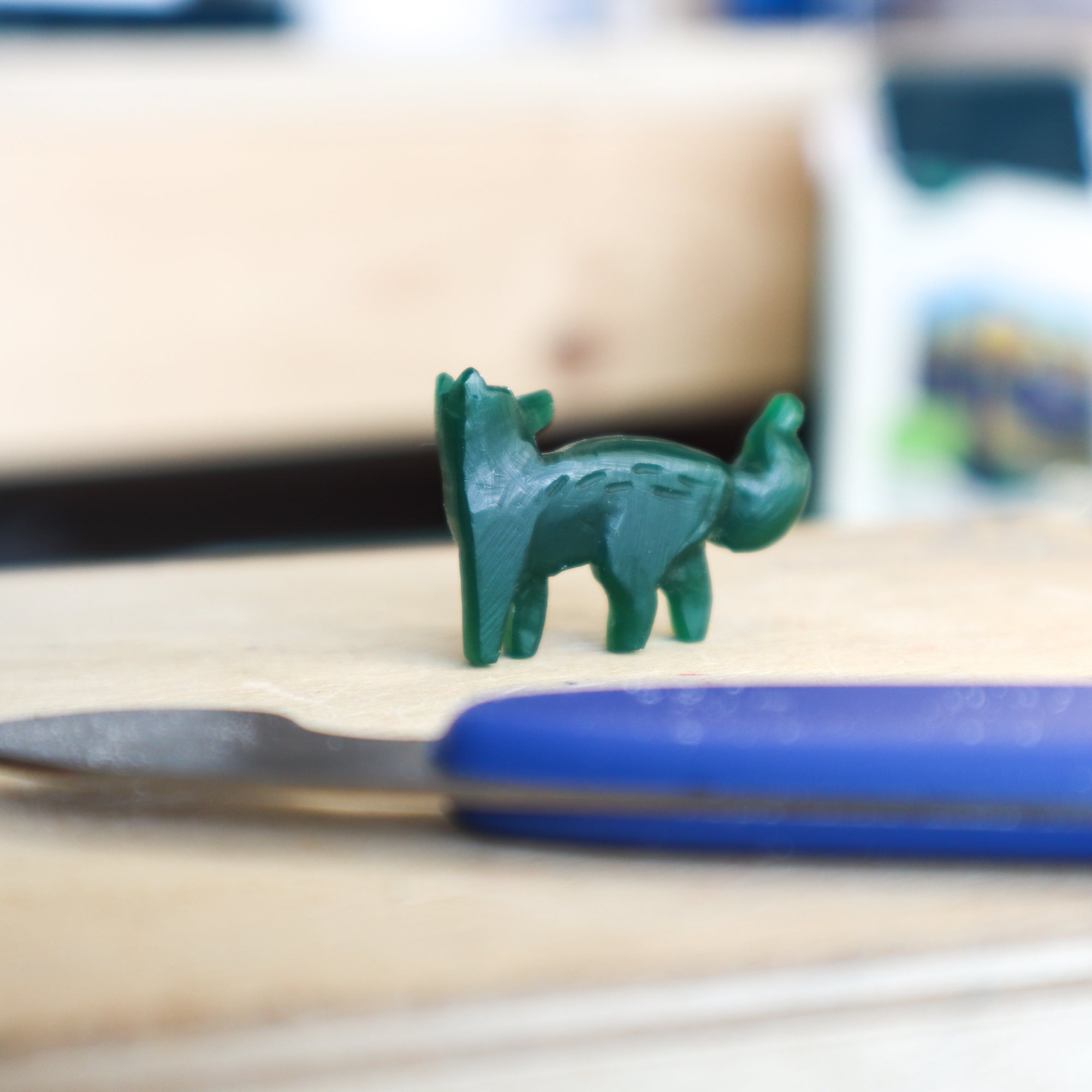 A small green fox figurine stands on wood with a blue-handled sculpting tool in front, evoking the artistry behind Papio Presss handmade recycled silver jewelry, including their Silver Fox Pendant. The softly blurred background adds charm.