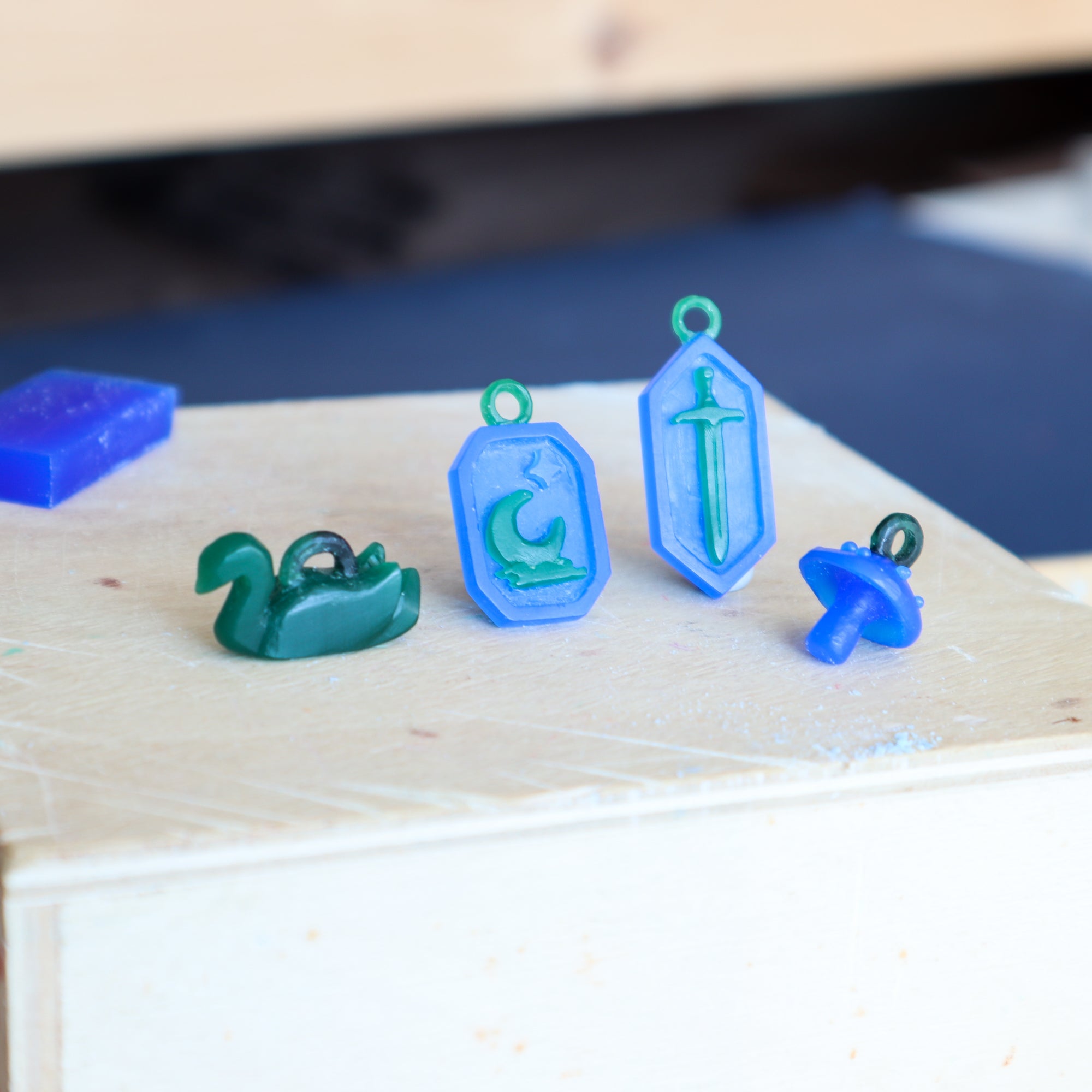 Green and blue plastic charms sit on a light wooden surface, including a green swan, mushroom, and two blue pendants—one featuring a carved sword design inspired by the Papio Press Silver Warrior Pendant. A blurry blue object appears in the background.