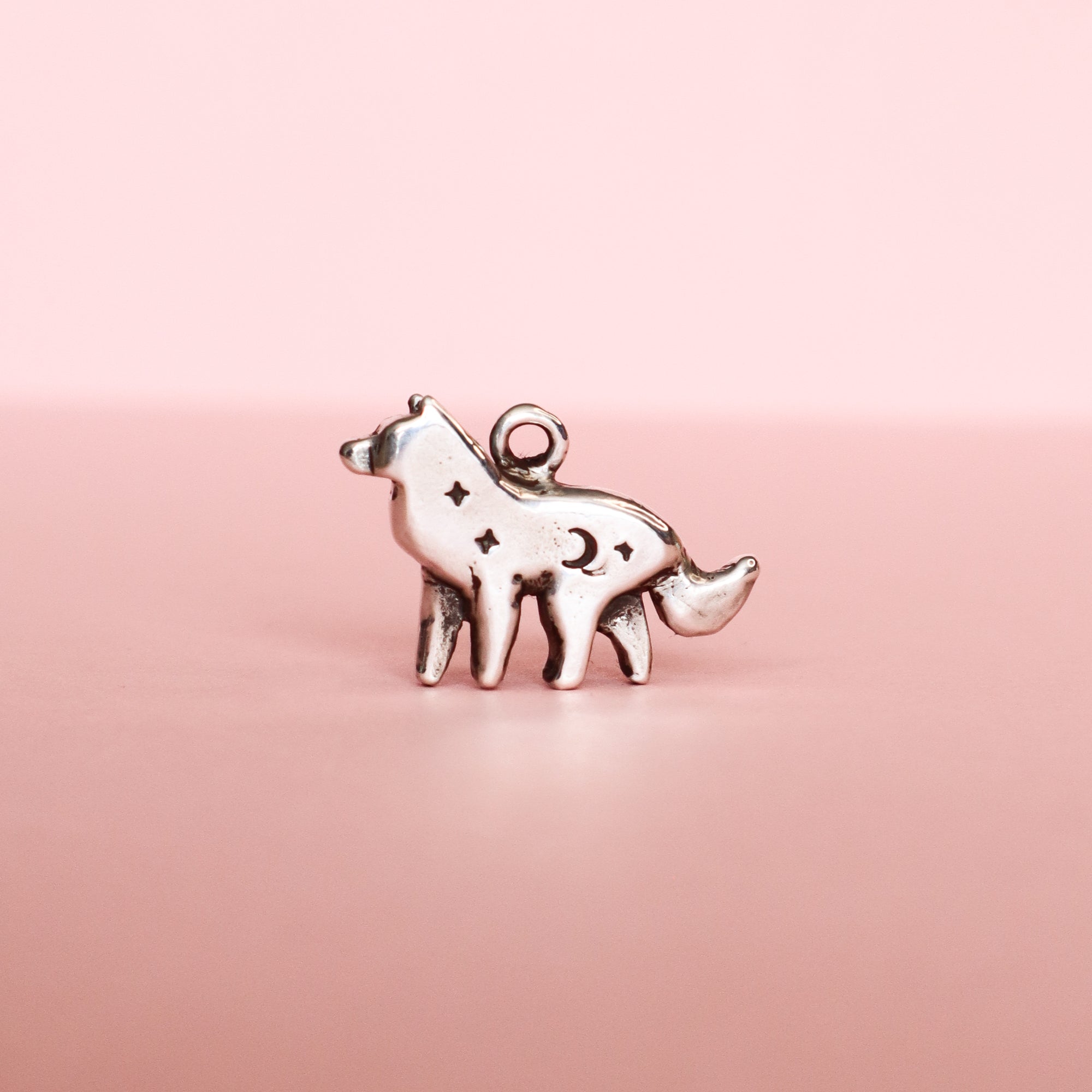 The Silver Lunar Wolf Pendant by Papio Press is a sterling silver necklace with a wolf-shaped pendant featuring cut-out stars and a crescent moon, displayed on a soft pink background.