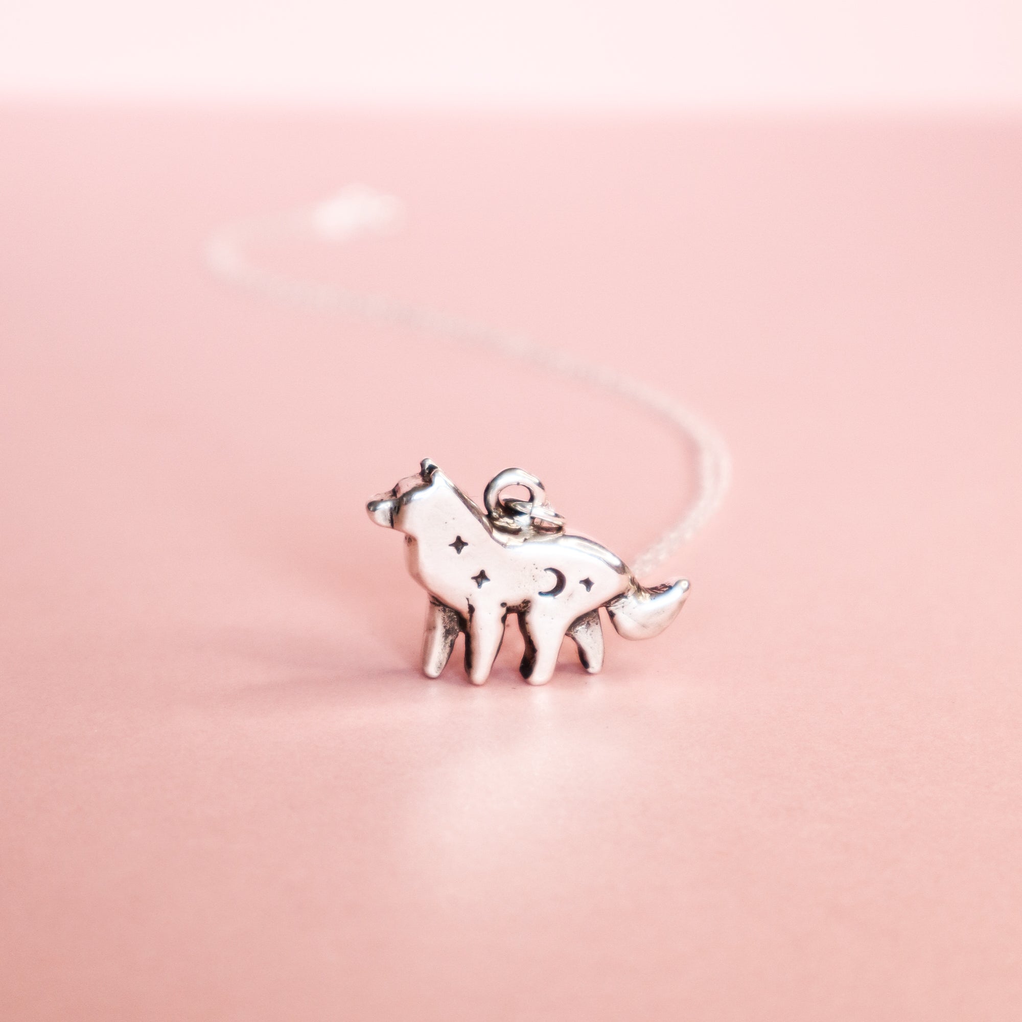The Papio Press Silver Lunar Wolf Pendant, a sterling silver necklace with star and moon engravings, is showcased on a soft pink background.