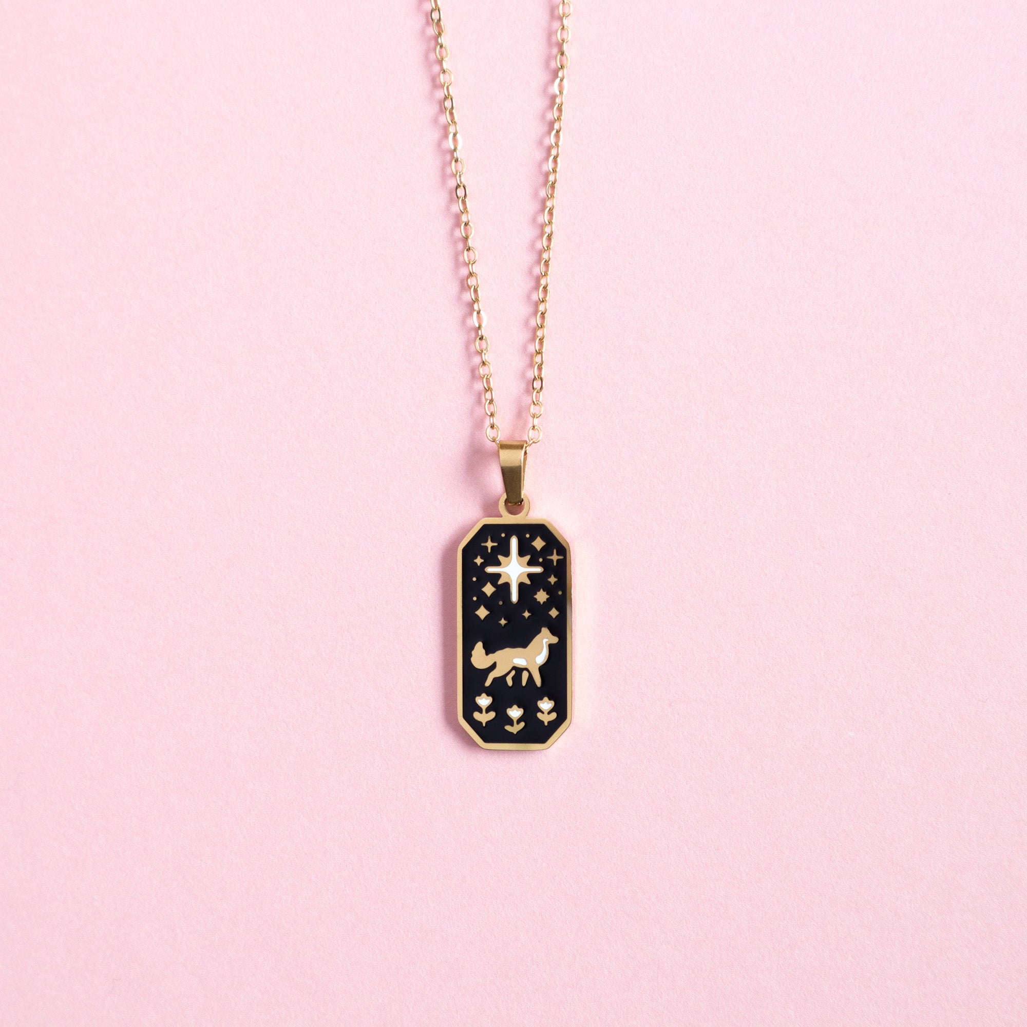 The Papio Press Wolf & Star Enamel Necklace features a gold stainless steel rectangular pendant with a cutout wolf, stars, and a large star, shown against a pink background.
