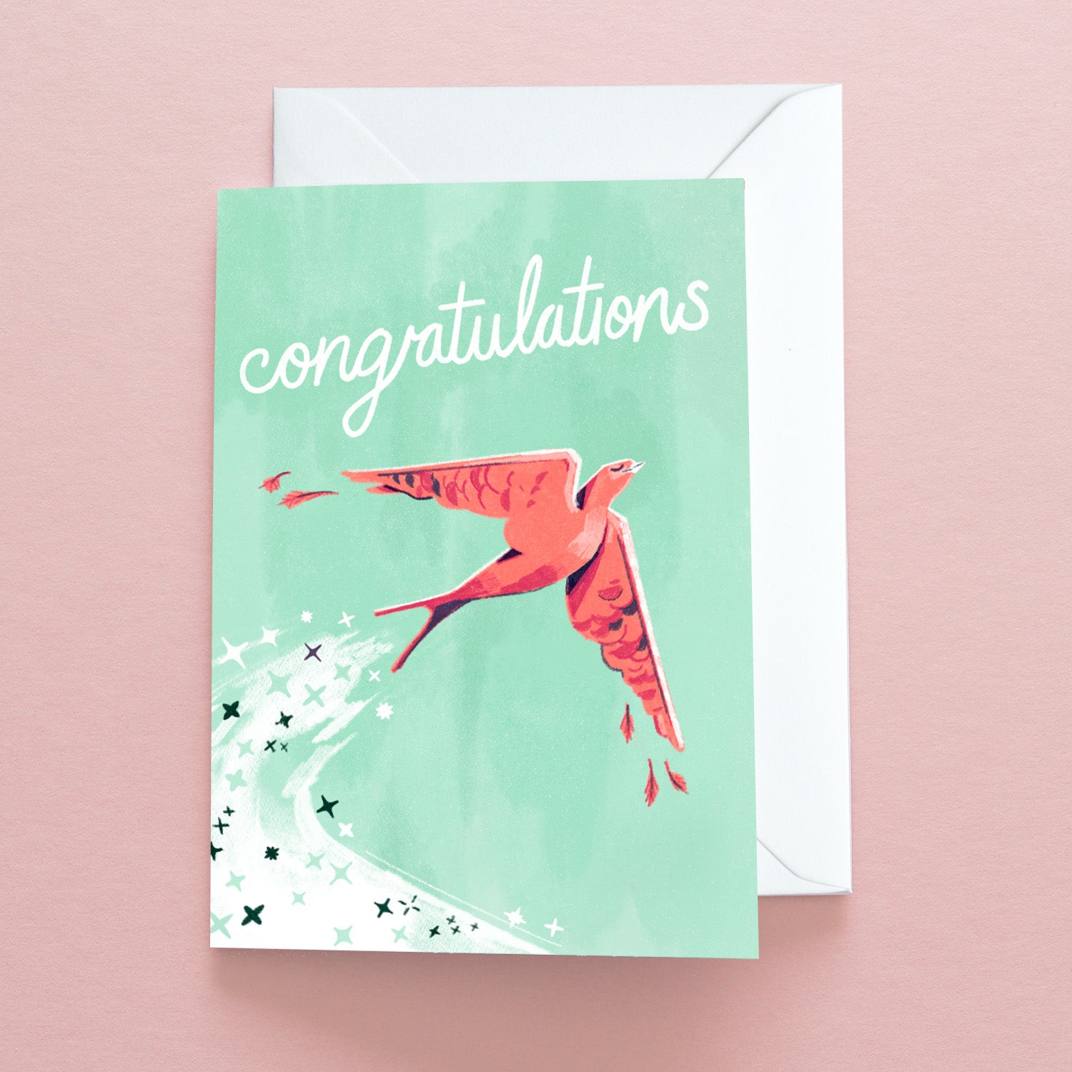 The Congratulations Flight Greetings Card by Papio Press features congratulations in white script above a pink bird in flight with a sparkling trail. Printed on recycled stock, it comes with a white envelope against a pale pink background.