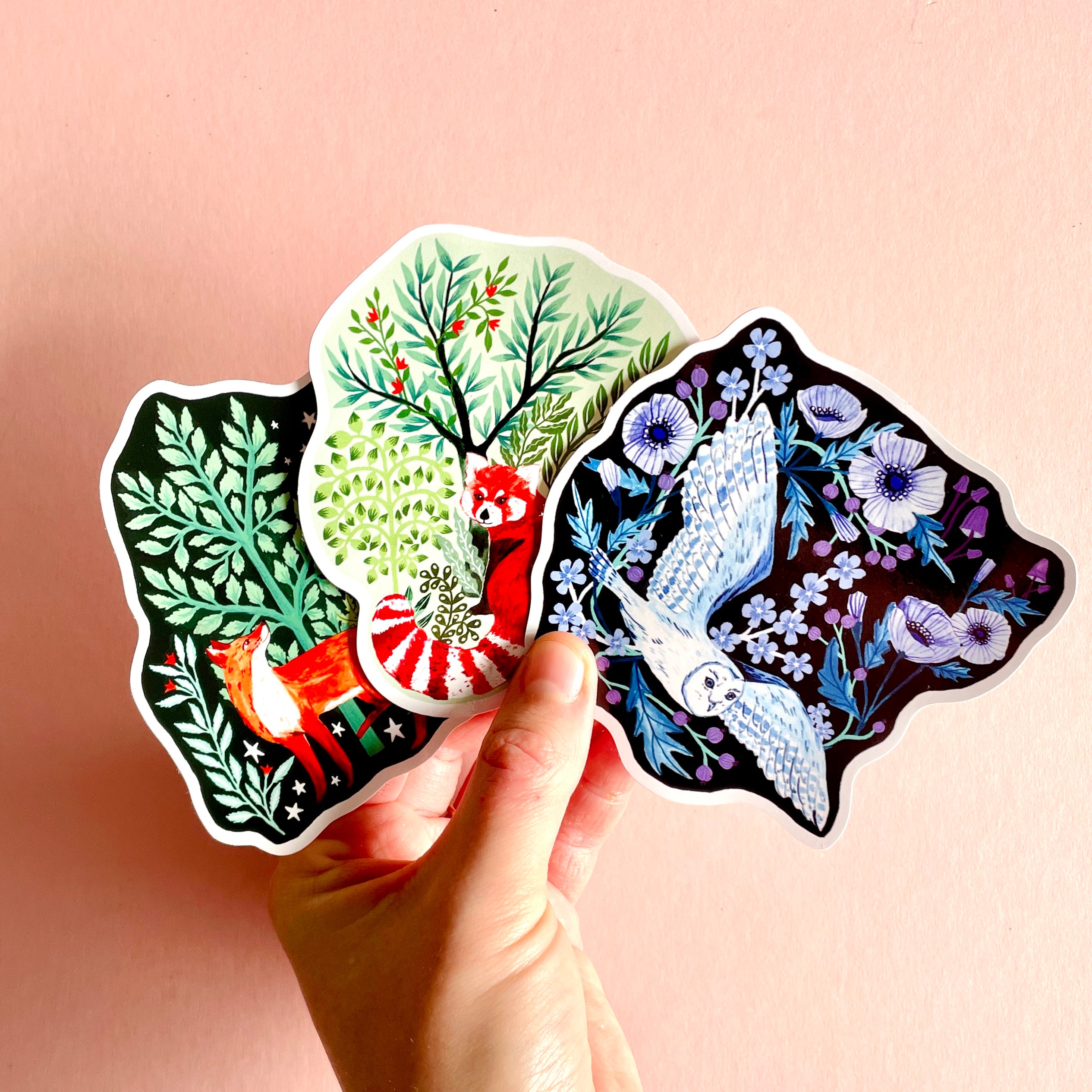 A hand displays Papio Presss Mystery Pack of Vinyl Stickers—featuring a fox in green foliage, a red panda in leafy branches, and a white owl with blue flowers—all made of durable vinyl, set against a pink background.