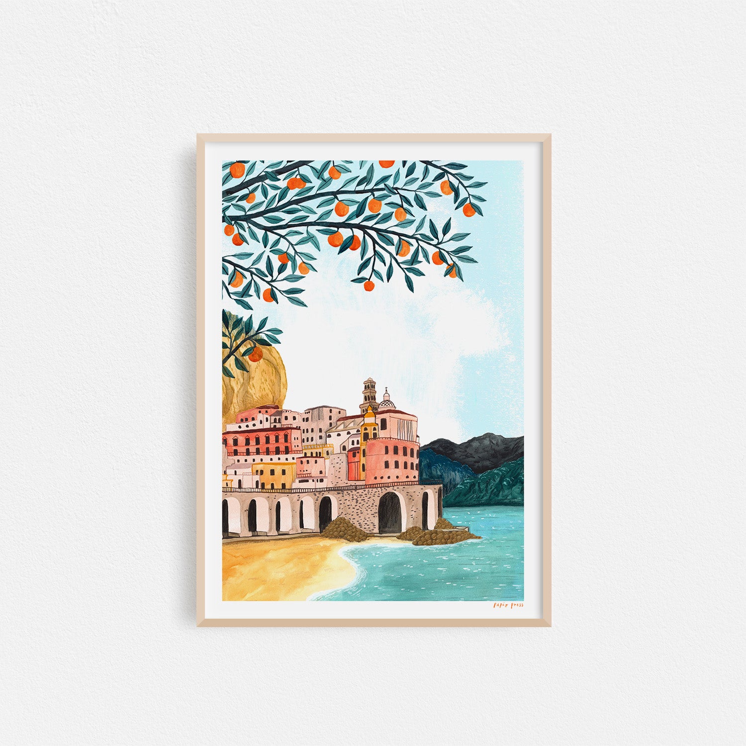 The Amalfi Coast Artists Print by Papio Press features a coastal town with pastel buildings, an orange tree arching over the top left, mountains, and a light blue sky in an inkjet print on archival paper.