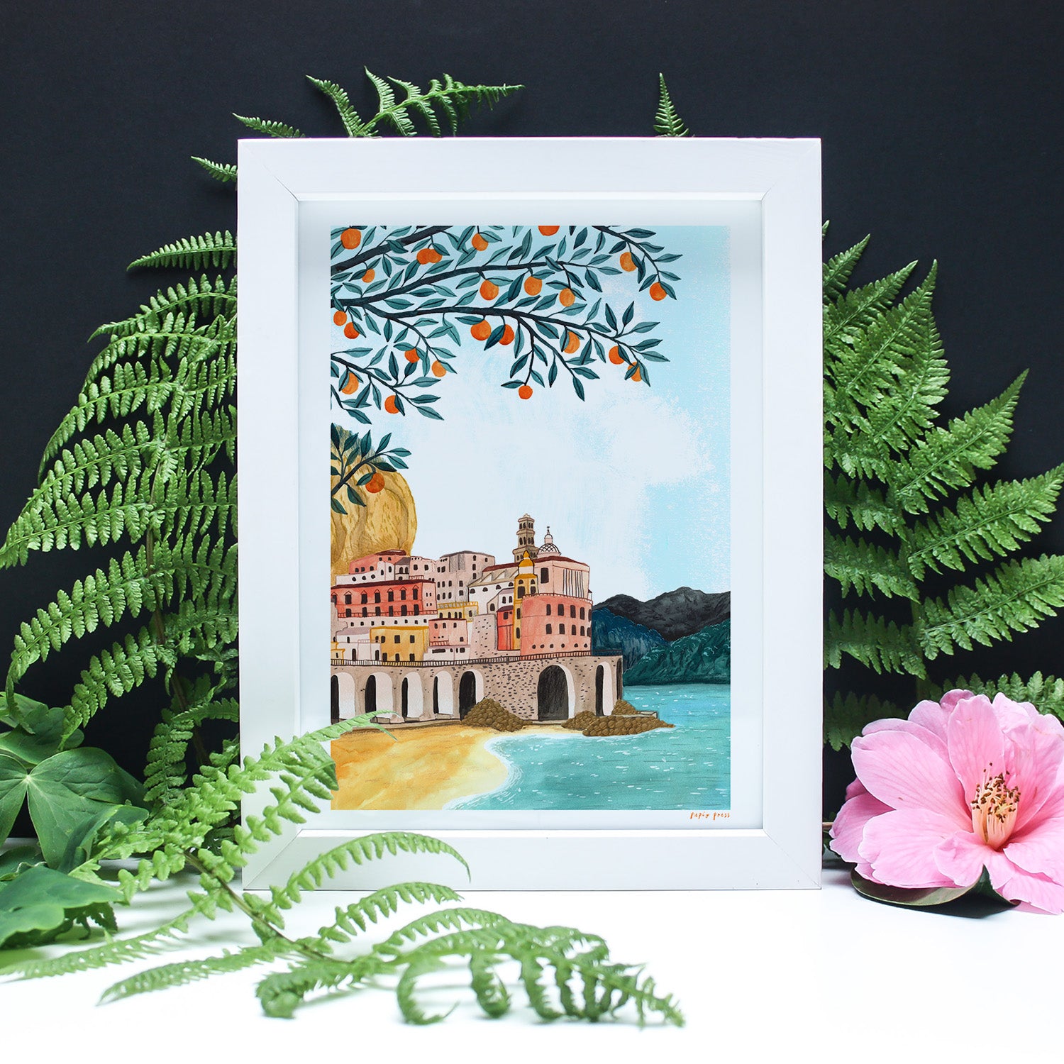 The Amalfi Coast Artists Print by Papio Press is a framed inkjet print of a vibrant coastal town, mountains, and orange tree branches, surrounded by ferns and hibiscus on archival satin paper with a black background.