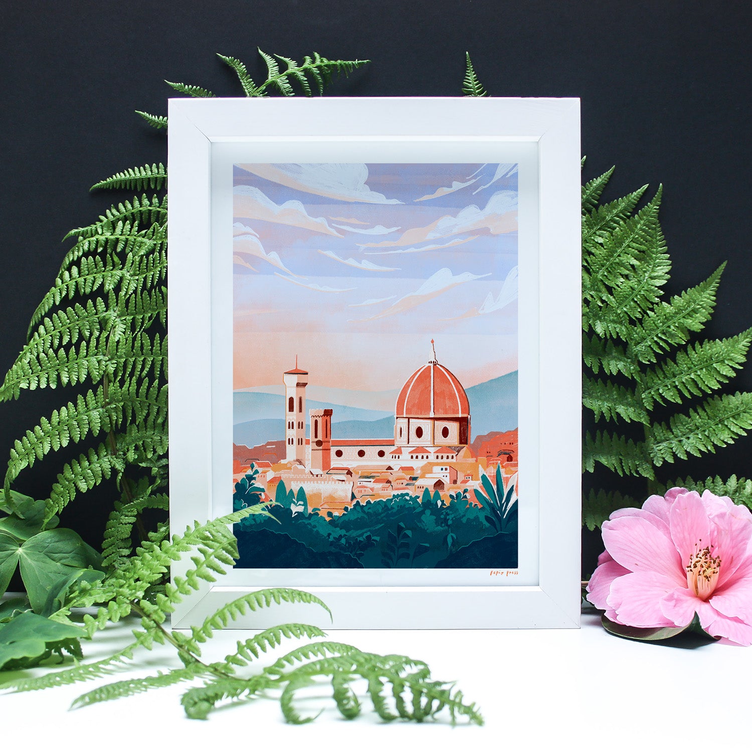 A framed painting of a cityscape with a prominent domed building, surrounded by green ferns and a pink hibiscus flower, set against a dark background.