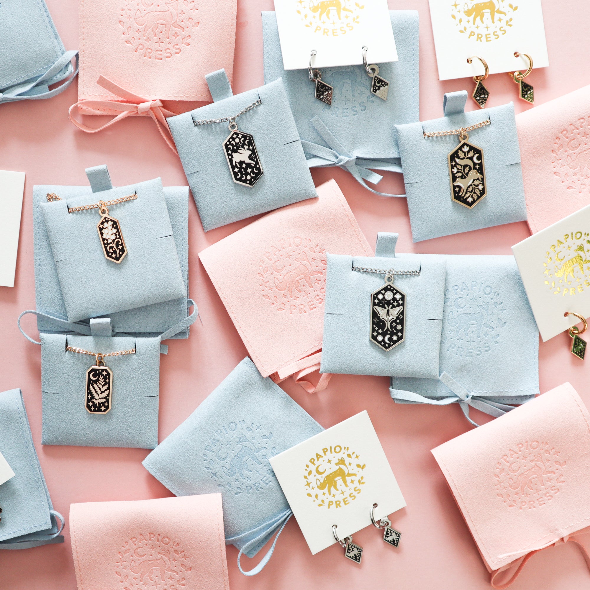 A selection of Papio Press necklaces and earrings on a pink background.