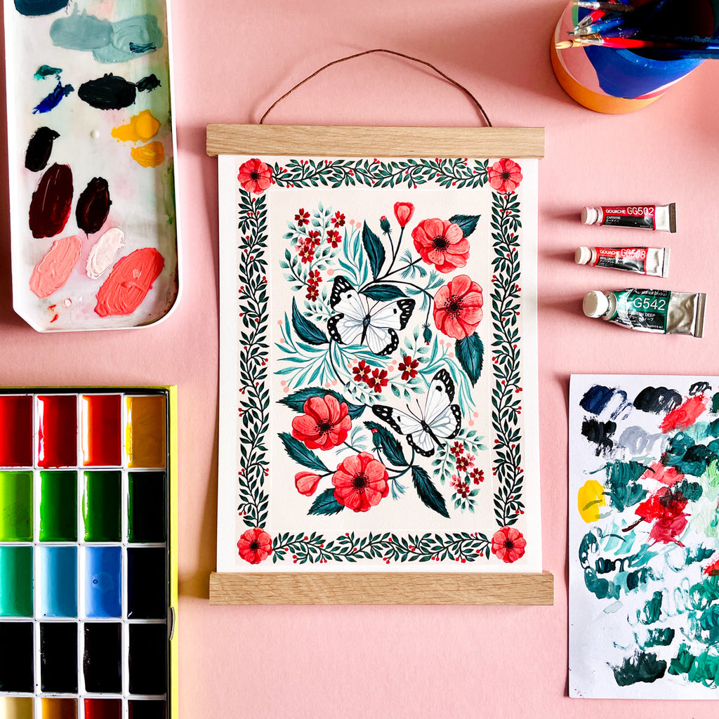 A photograph of a framed floral Butterly print on a pink background. The print is surrounded by gouache paints, paint brushes and paint smears.
