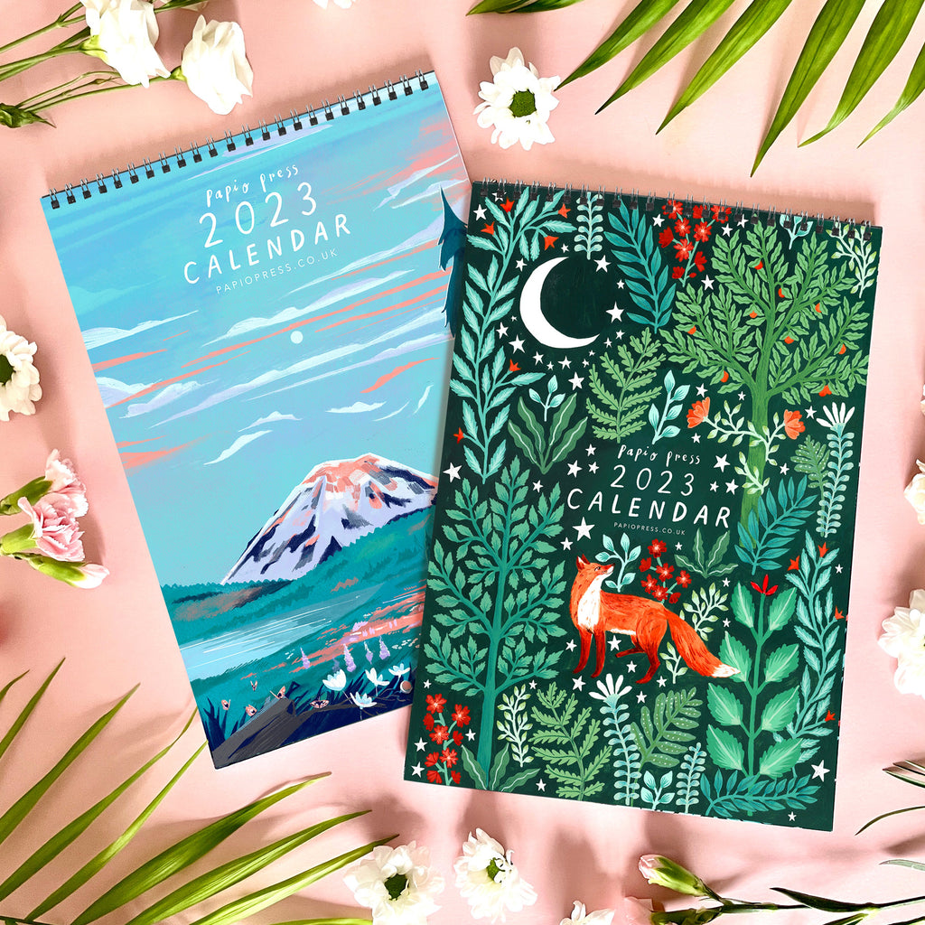 A photograph of two 2023 Papio Press calendars. One calendar features an illustration of Mt. Rainier and the other features an illustration of a woodland fox surrounded by trees, foliage and the moon.