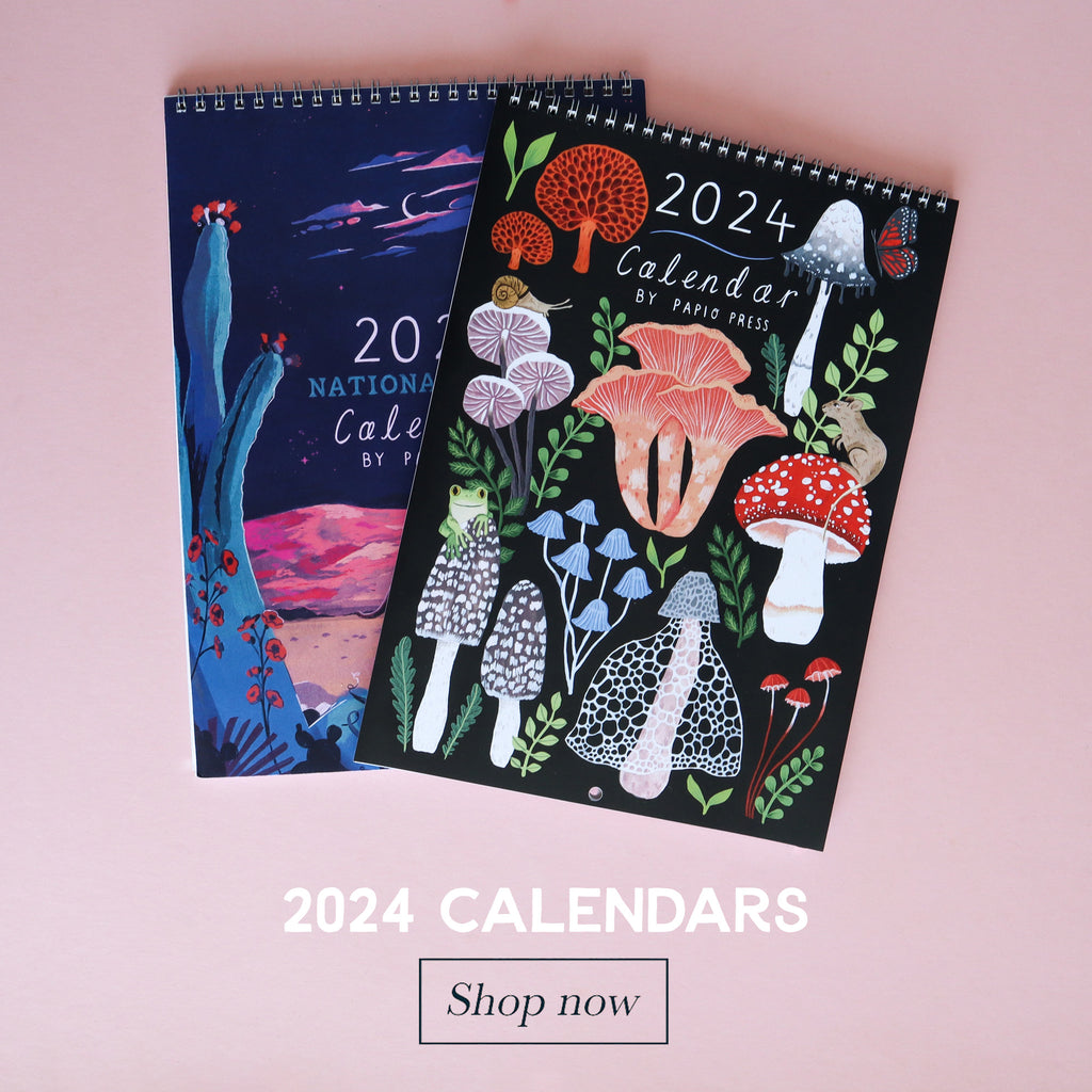 A photograph of two 2024 calendars on a pink background. One calendar features gouache paintings of some mushrooms and foliage, the other calendar is an illustration of a desert scene featuring some cacti.