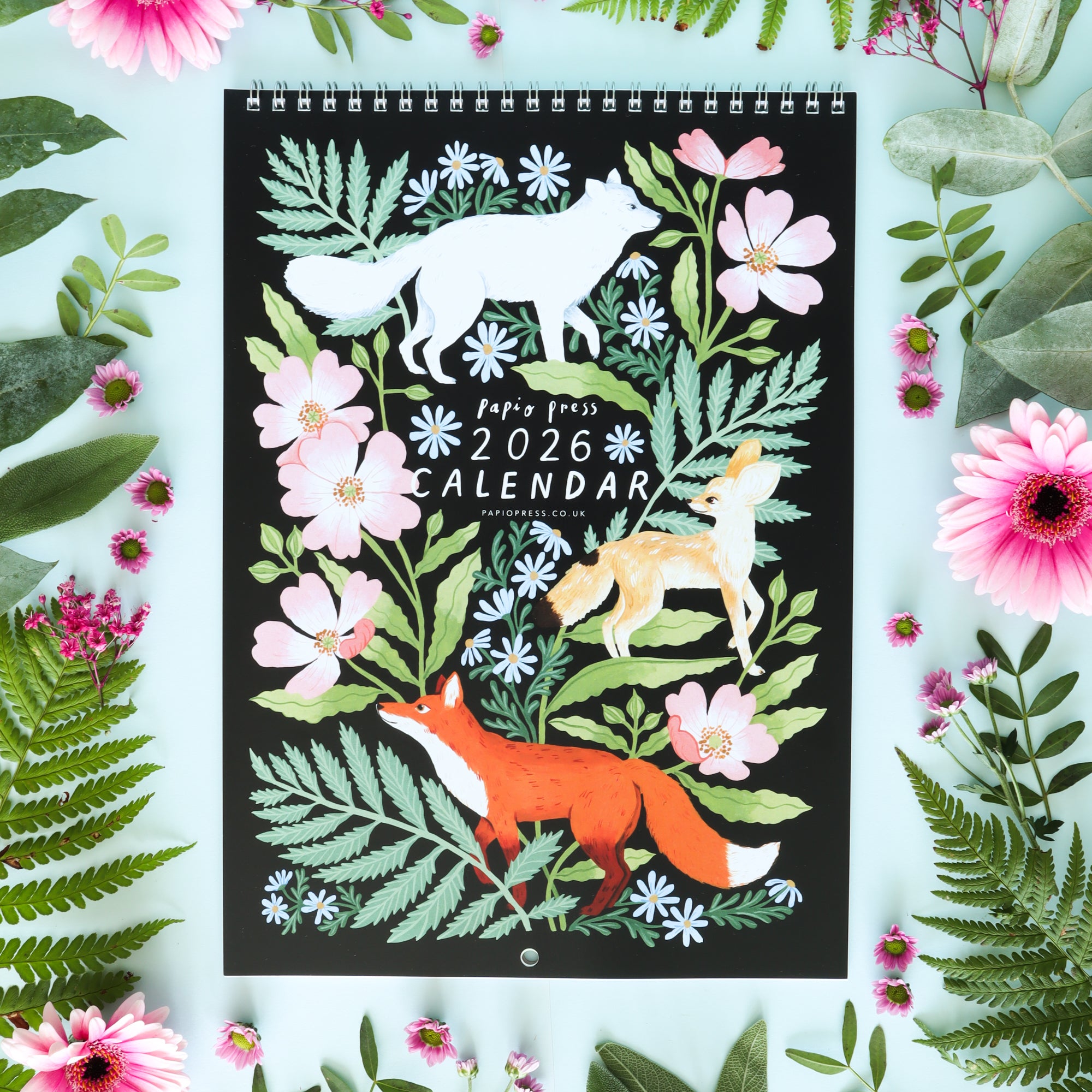 The 2026 Papio Press Florals & Animals Calendar features illustrated foxes with pink flowers on a black background, surrounded by ferns and florals on blue.
