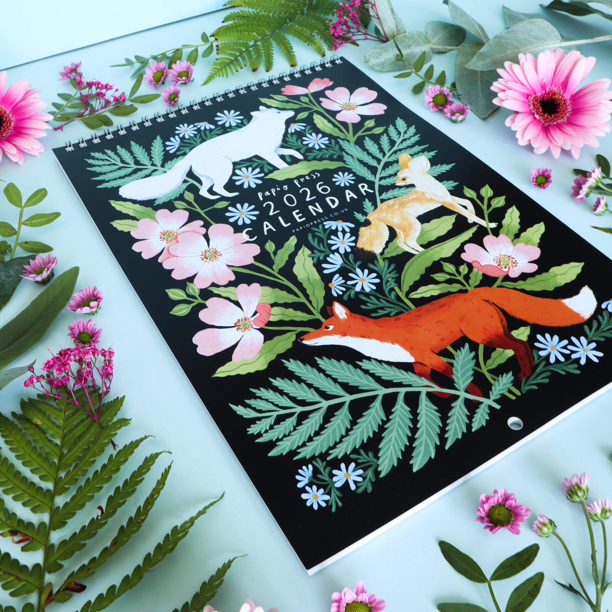 The 2026 Papio Press Florals & Animals Calendar by Papio Press features a black cover with foxes, pink flowers, and green leaves, inspired by their prints, displayed among real ferns and pink blossoms on a light blue background.