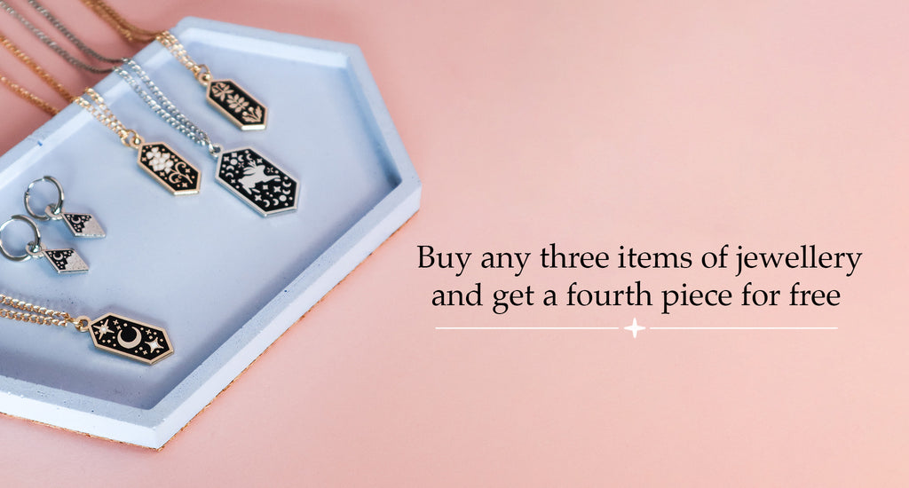 A blue concrete tray featuring four necklaces and a pair of silver earrings. The tray is on a pink background. It features text that says 'Buy any three items of jewellery and get a fourth piece for free'. 