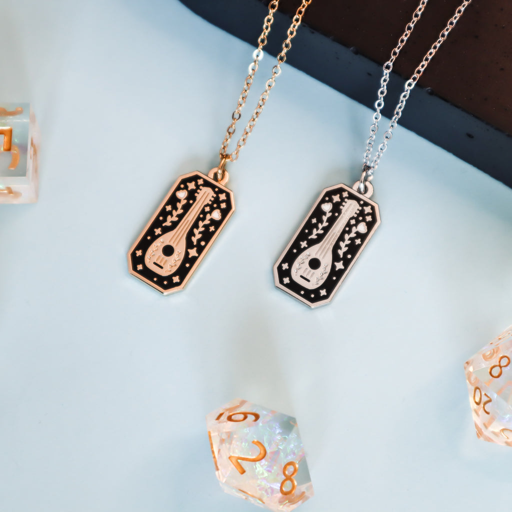 Two bard necklace, one silver and one gold on a light blue background. The necklaces are rectangular and feature a lute, flowers and stars with black and white enamel details.
