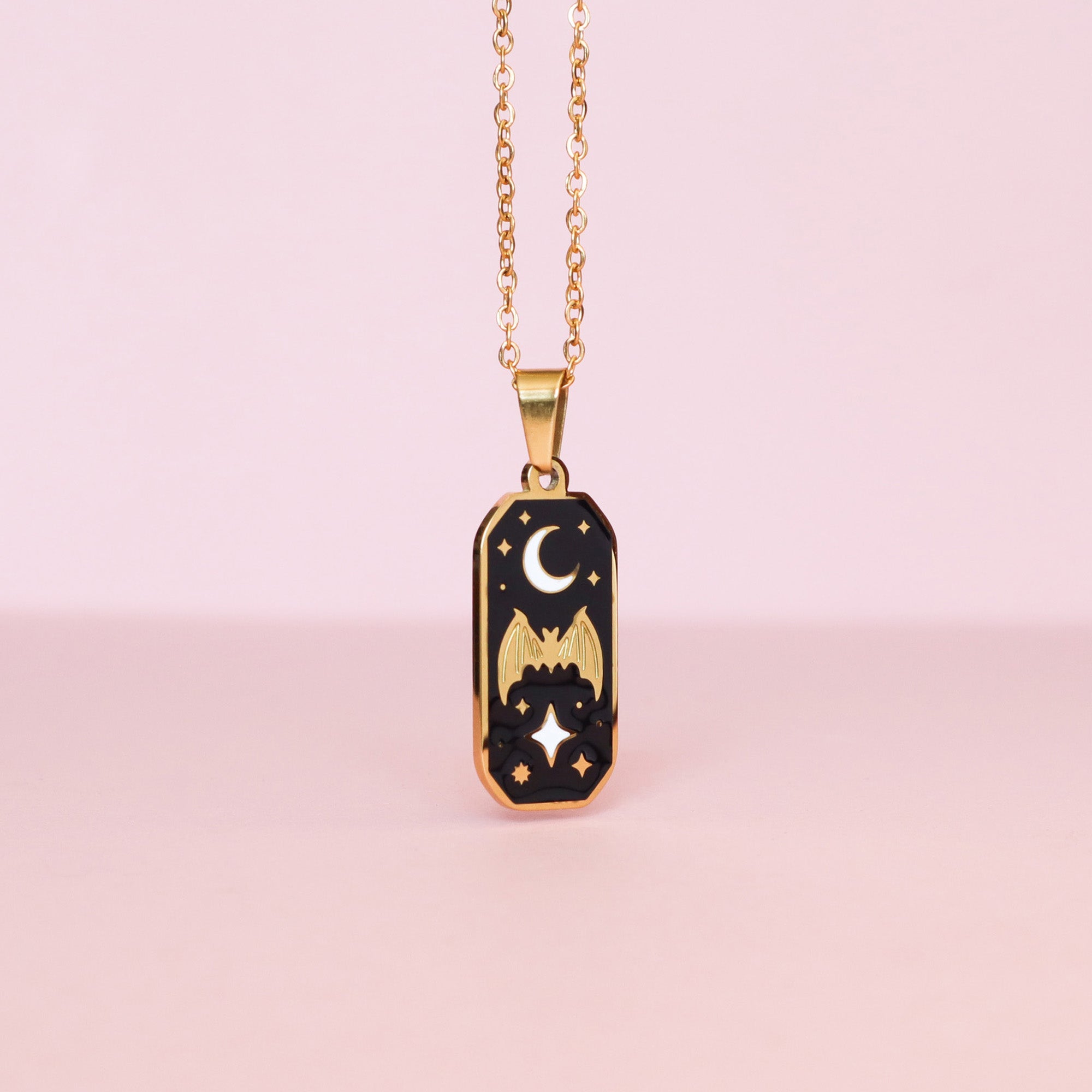 The Bat & Moon Enamel Necklace by Papio Press features a rectangular black pendant with gold bat, moon, and star details on stainless steel, shown against a soft pink background.