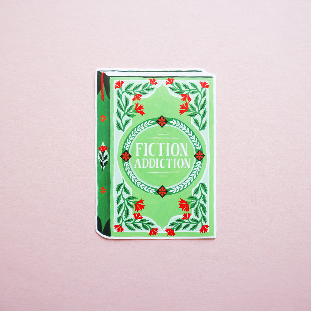 A picture of a book sticker that says 'Fiction Addiction'. The photo is taken on a pink background.