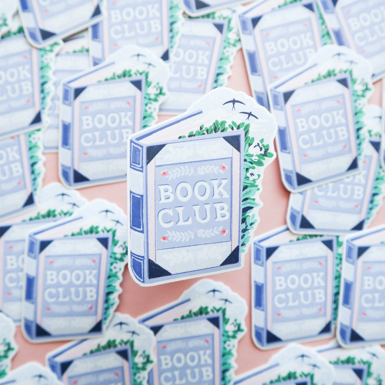 Papio Press’s Book Club Book Sticker features a blue book with leafy decorations, pink dots, and flying birds. Made of durable vinyl, its perfect for any book lover and comes scattered on a light pink background.