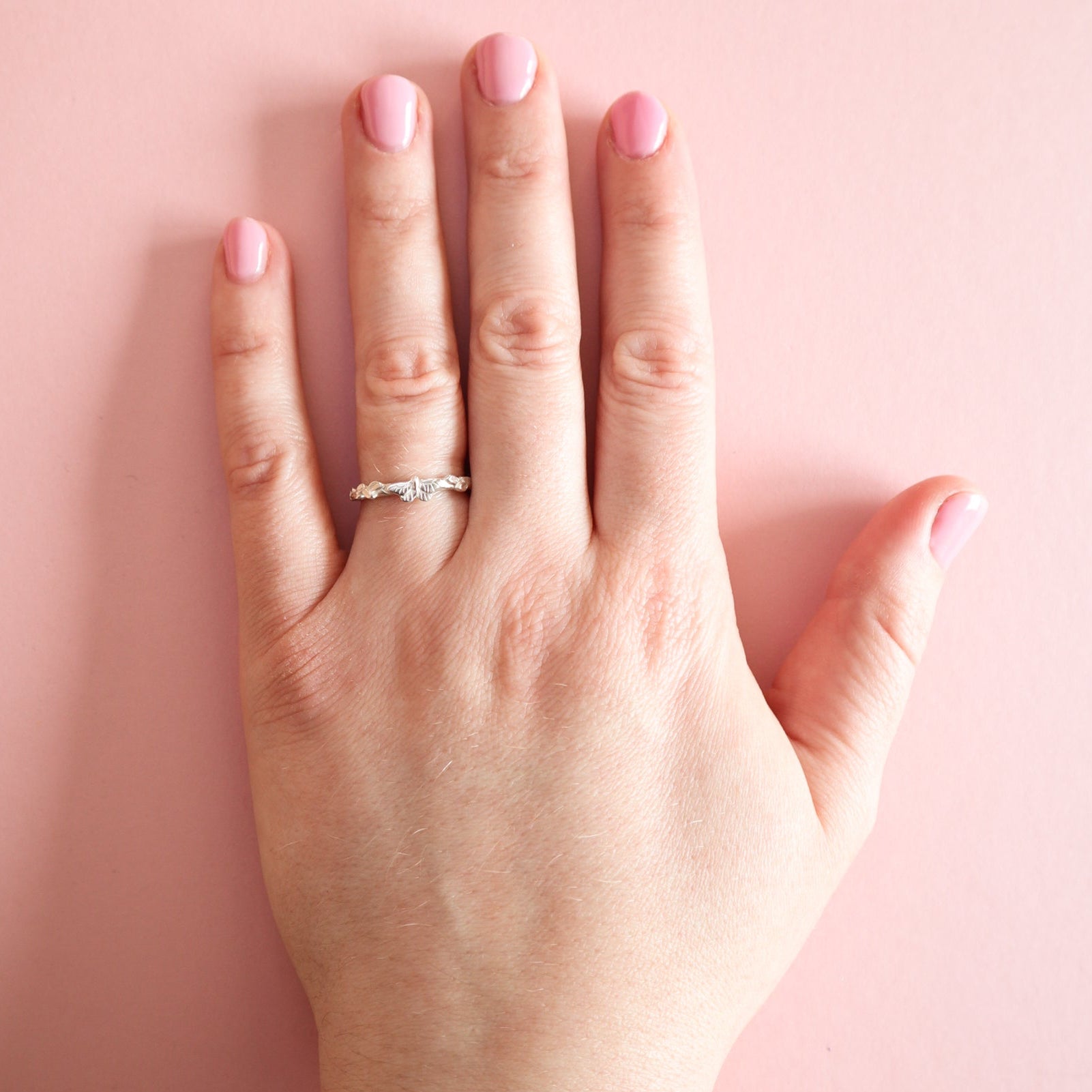 A hand wearing a silver butterfly ring on a light pink background. 