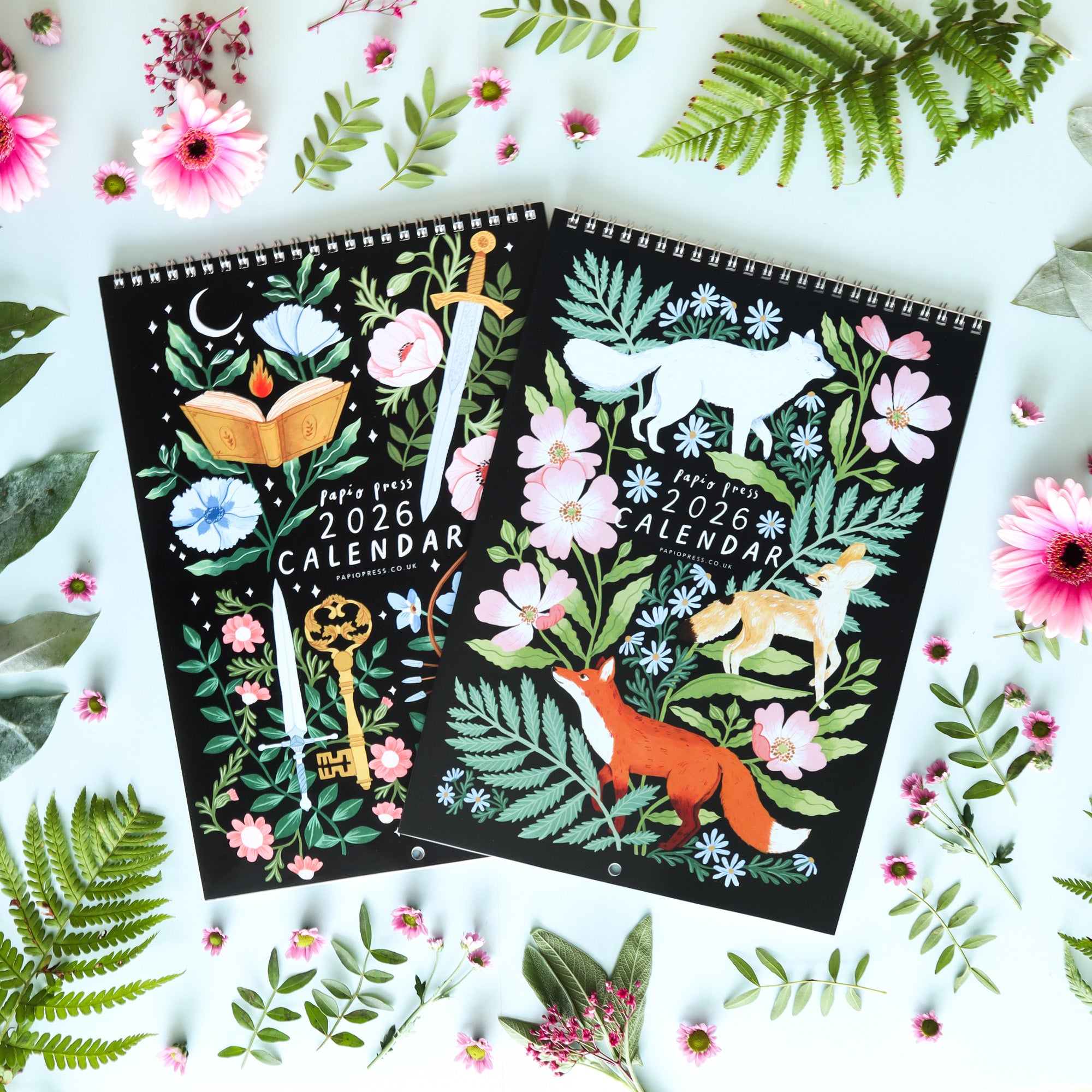 A Papio Press gift set of two 2026 calendars rests amid green leaves and pink flowers. One cover shows a some florals and foxes, the other displays an open book, sword, and key with blue and pink blooms on black.