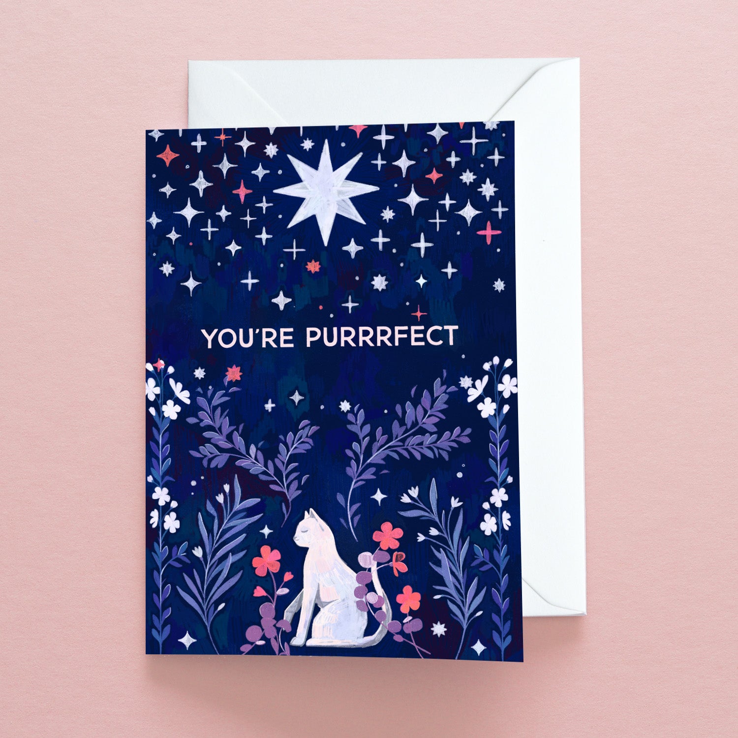 The “Youre Purrrfect Greetings Card” by Papio Press features a white cat among florals under starry skies on dark blue. Printed on recycled stock, it reads YOURE PURRRFECT and is shown resting on a pale pink surface.