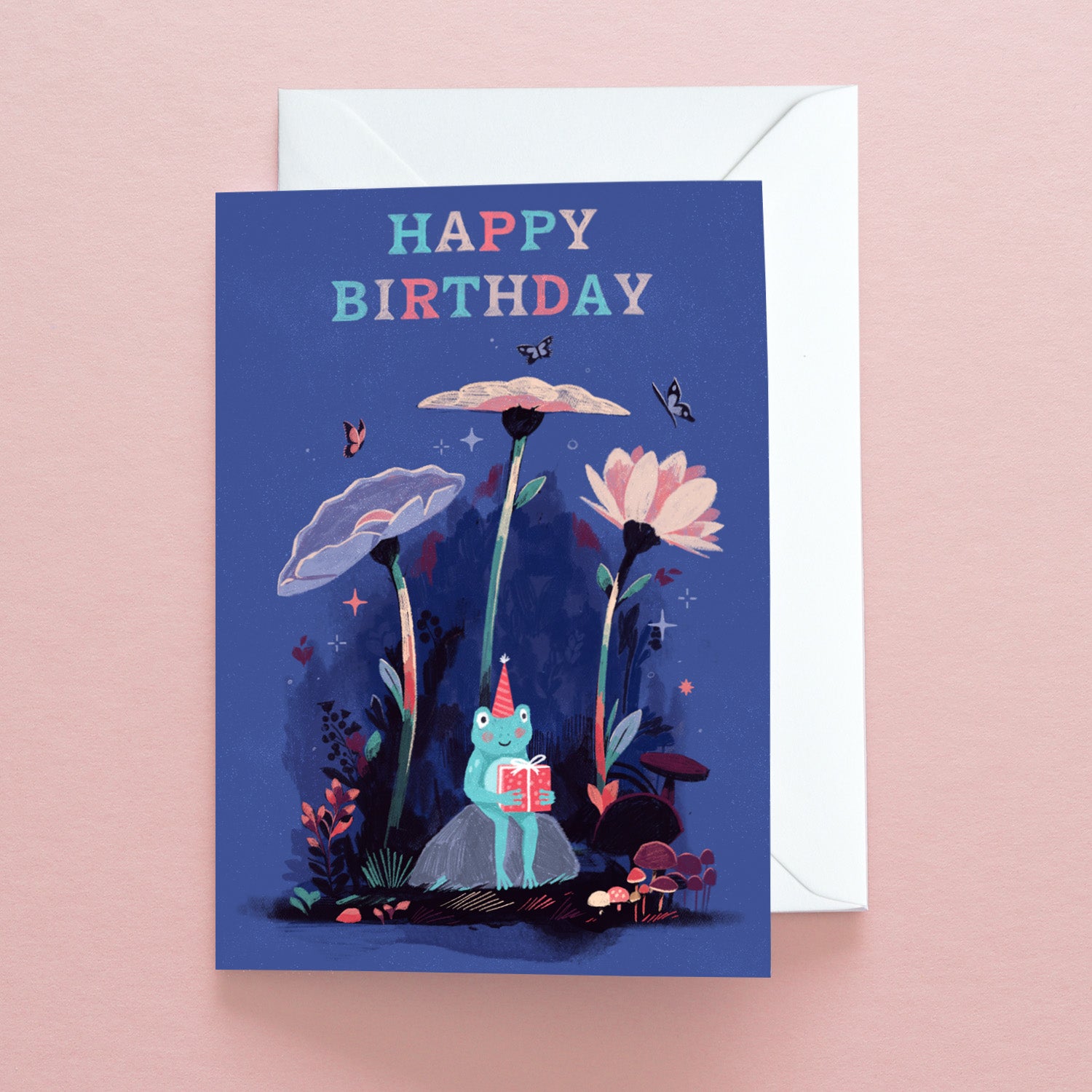 The Papio Press Happy Birthday Frog Greetings Card features a frog in a party hat holding a present, with colorful HAPPY BIRTHDAY text above. Surrounded by mushrooms, flowers, and butterflies, its printed on recycled stock.
