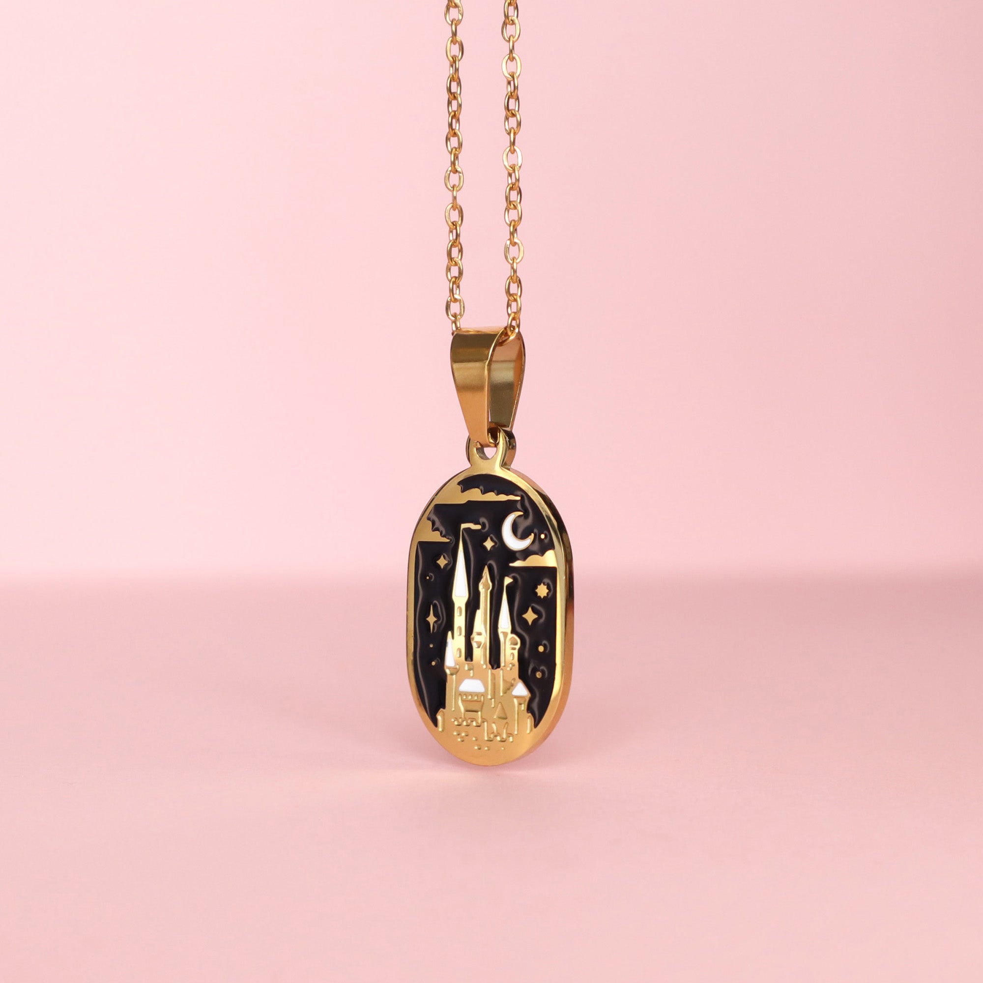 The Moonlit Castle Enamel Necklace by Papio Press features a gold oval charm with a moonlit castle, stars, and crescent moon on black enamel over stainless steel, displayed on a soft pink backdrop.