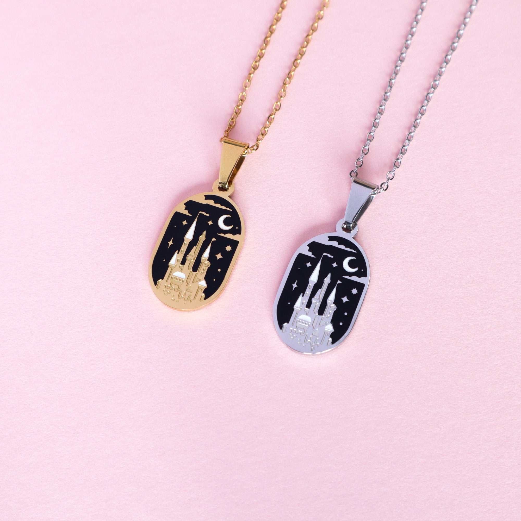 Two Papio Press Moonlit Castle Enamel Necklaces, one gold-tone and one silver-tone stainless steel, each featuring a castle, stars, and a crescent moon on black enamel, displayed on a soft pink surface.