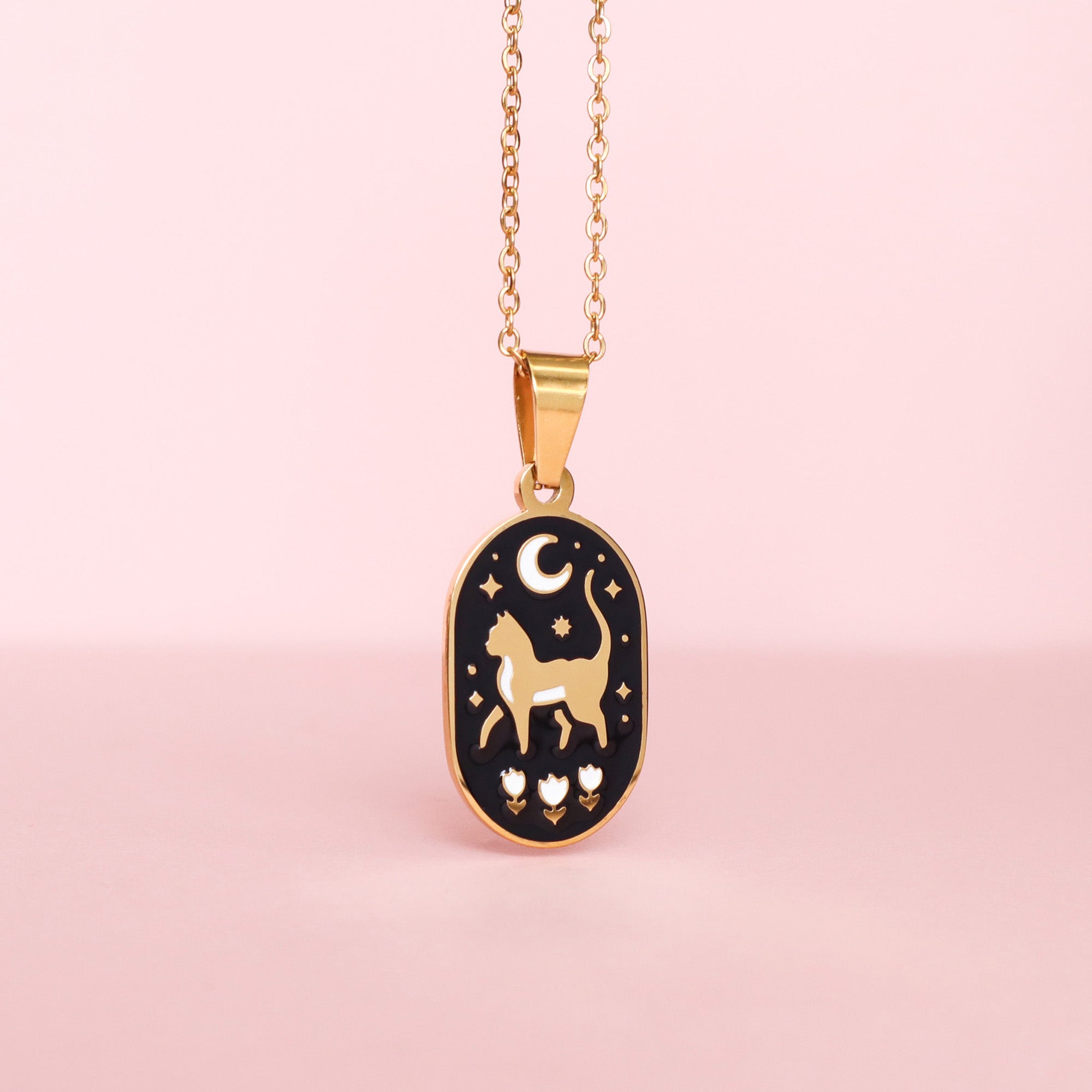 The Cat & Moon Enamel Necklace by Papio Press features a gold stainless steel chain with a black charm displaying a cat silhouette, crescent moon, stars, and three flowers on a pink background.