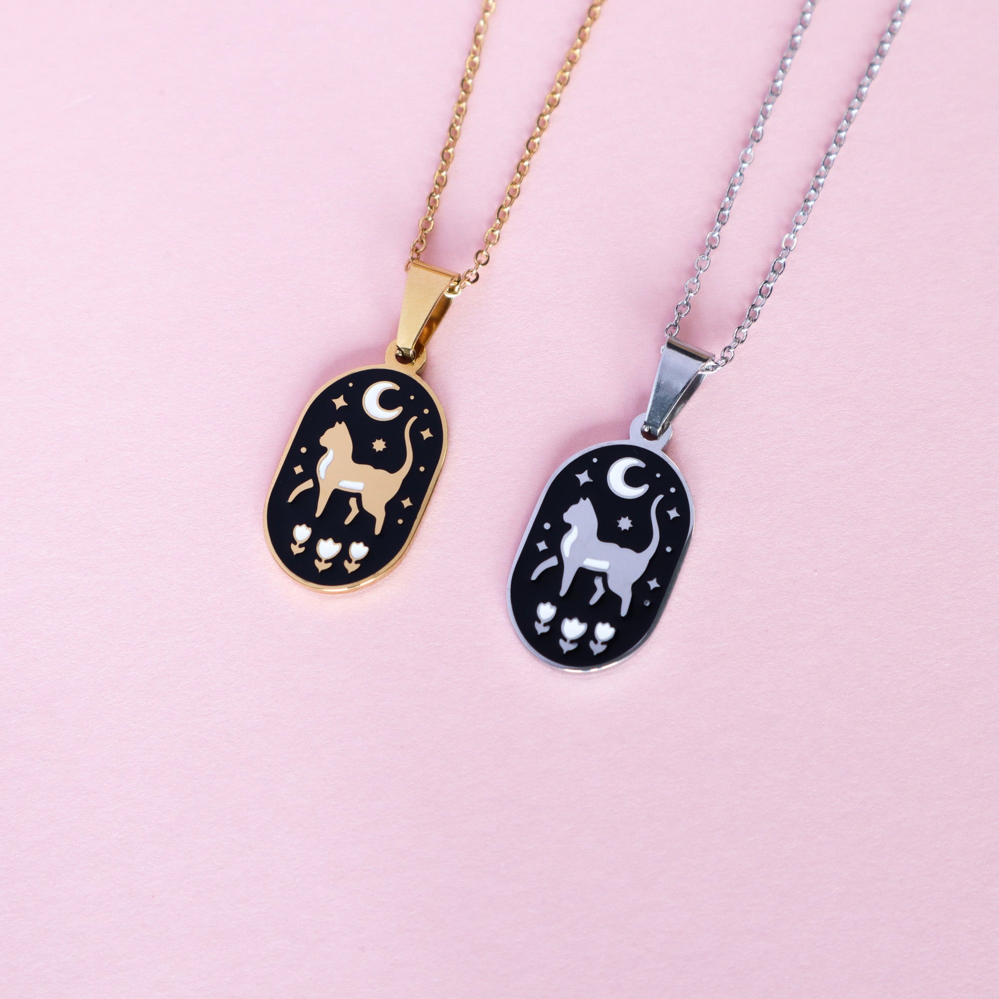 Papio Press Cat & Moon Enamel Necklaces: two oval pendants on stainless steel—one gold, one silver—each with a black background, crescent moon, cat, stars, and flowers. Displayed on a light pink surface.