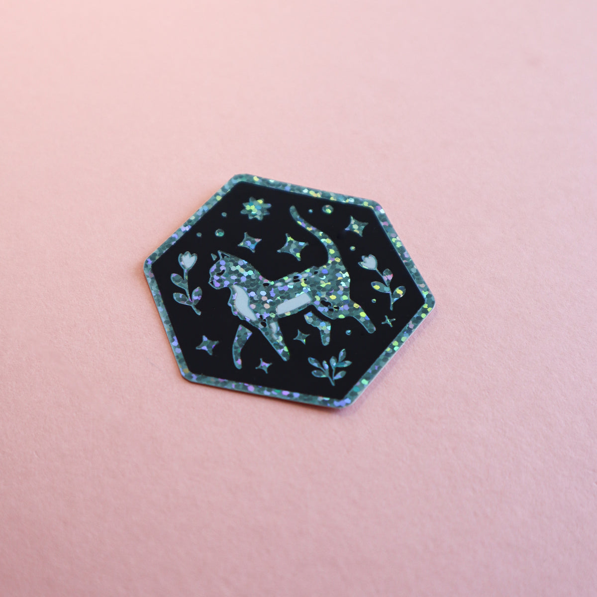 A hexagonal sticker featuring a glittery green cat surrounded by stars and leaves on a black background, placed on a solid pink surface.