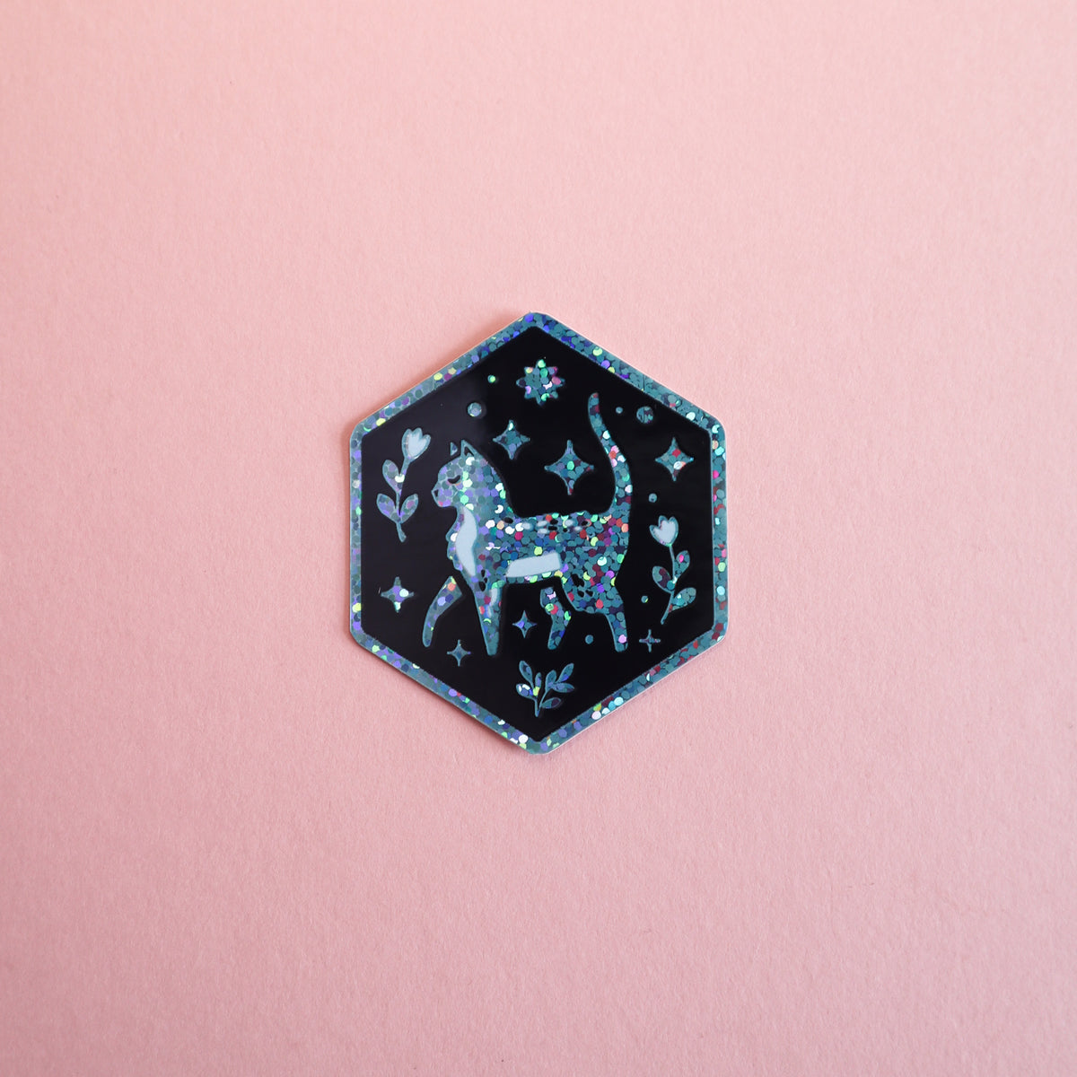 A hexagonal sticker featuring a glittery black cat surrounded by stars and flowers, set against a solid pink background.