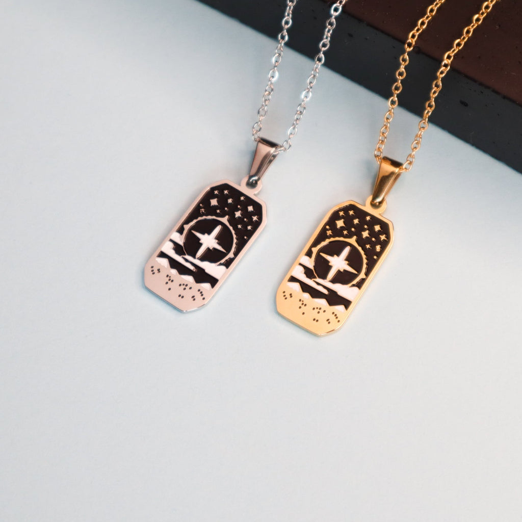 Two rectangular Cleric Pendant Enamel Necklaces by Papio Press—one silver, one gold—each featuring a bird, stars, and text at the bottom, displayed on a light blue surface. Ideal for tabletop game lovers.