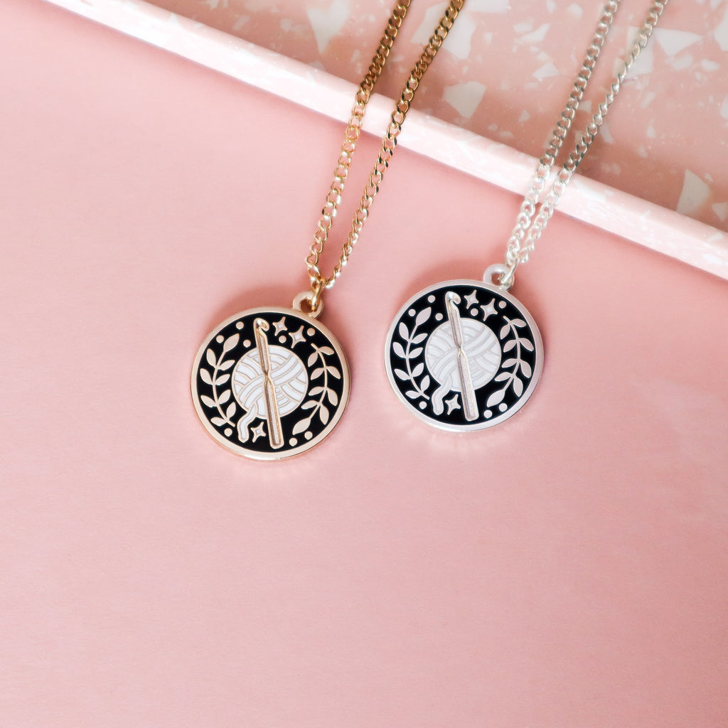 Two crochet necklaces, one gold and one silver. The design is circular and features a ball of yarn with a crochet hook surrounded by leaves and stars. It features black and white enamel details.