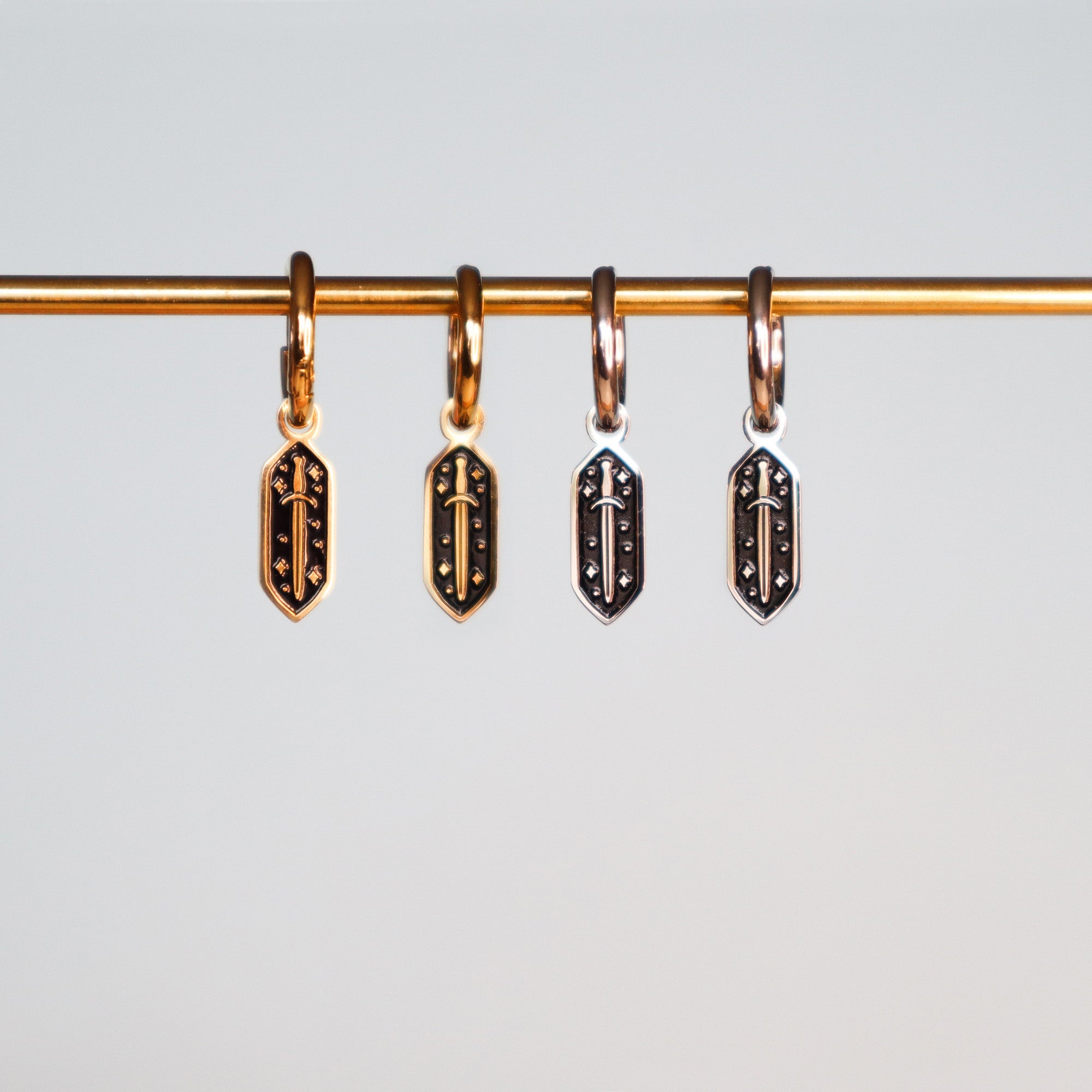 The Papio Press Mini Sword Hoop Earrings feature four shield-shaped charms in gold and silver tones, each with raised sword and star motifs on black—ideal for accenting stainless steel or enamel earrings.