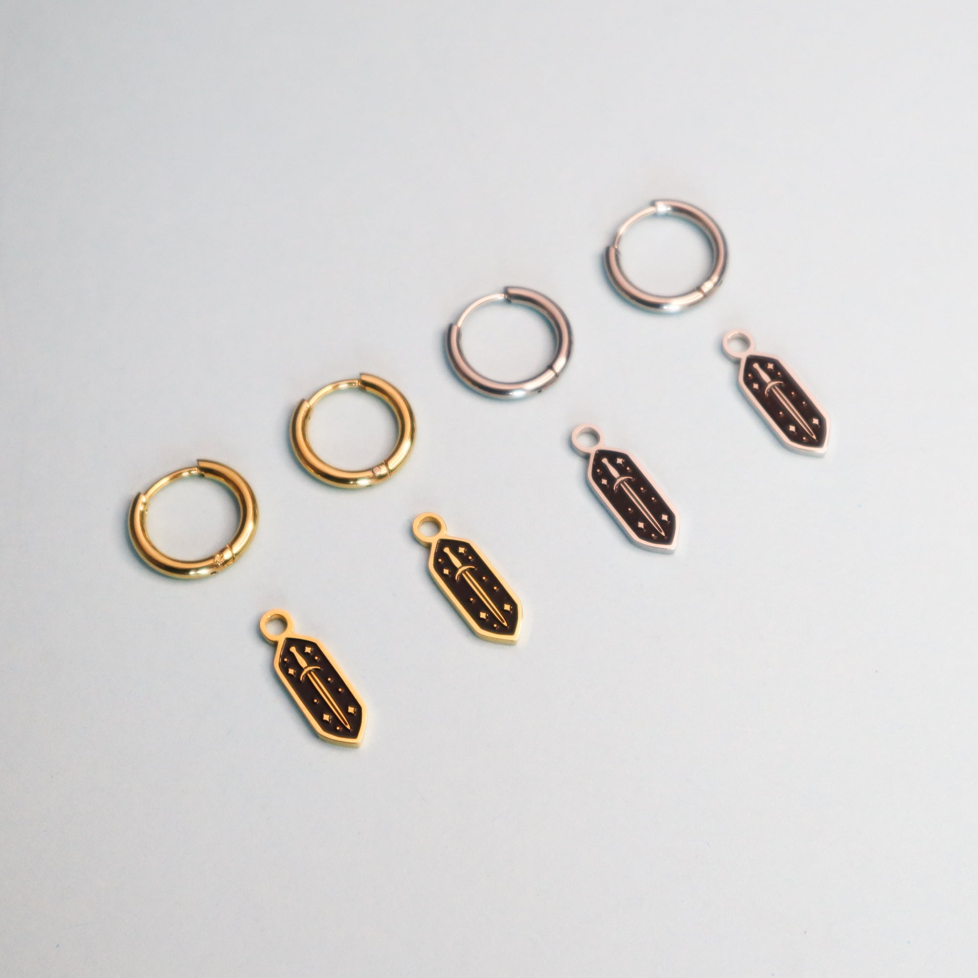 Four Papio Press Mini Sword Hoop Earrings—two gold, two silver—are displayed on a light surface, each paired with a matching feather-shaped charm in gold or silver placed just below.