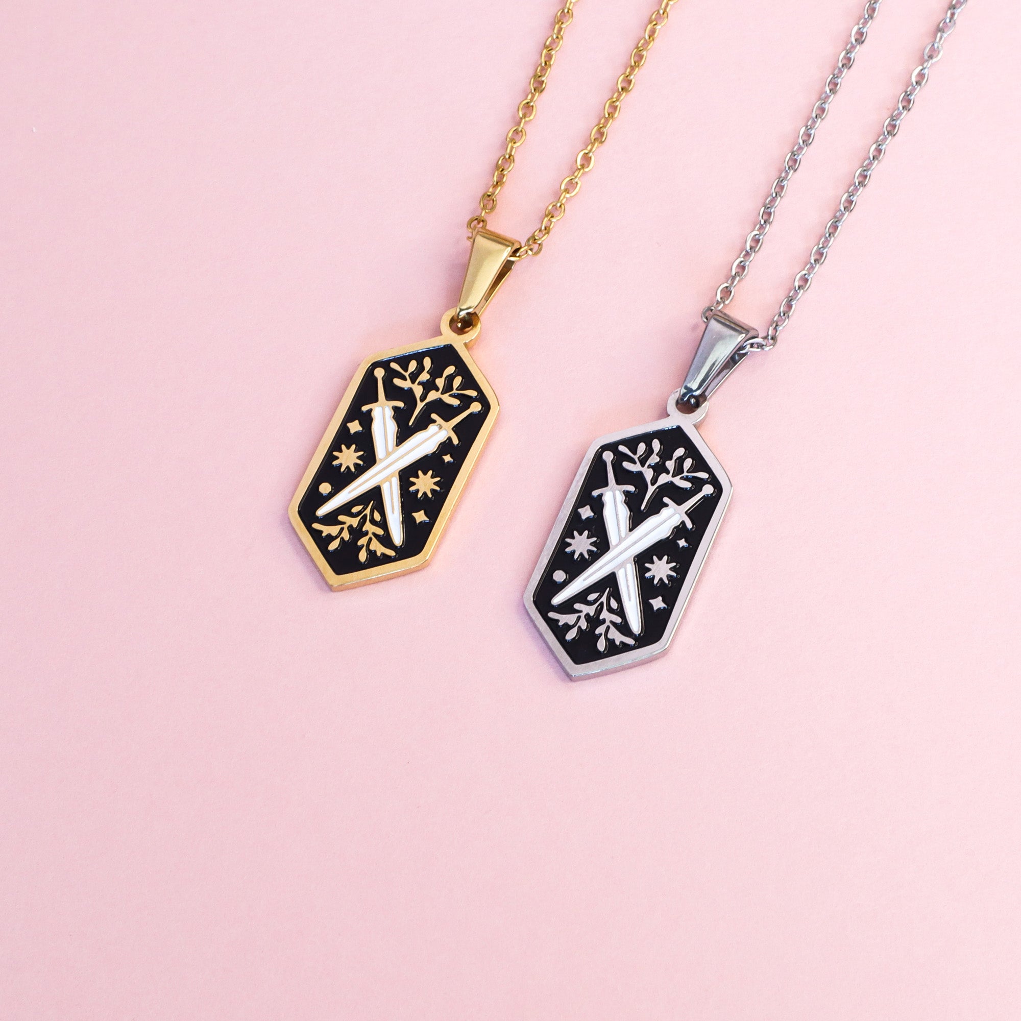 Two Papio Press necklaces in an elongated diamond shape. They feature two swords and leaves with white and balck enamel details on a pink back ground. 