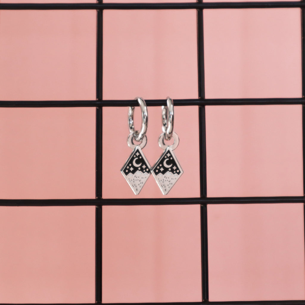 A pair of silver mountain earrings hanging on black wire with a pink background.