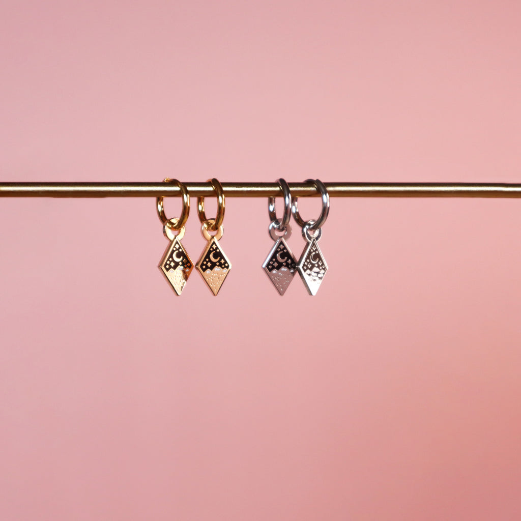 Two pairs of mountain earrings, one in silver and one in gold. They are diamond shaped and hang from a huggie hoop. They feature a mountain and moon with black and white enamel details.