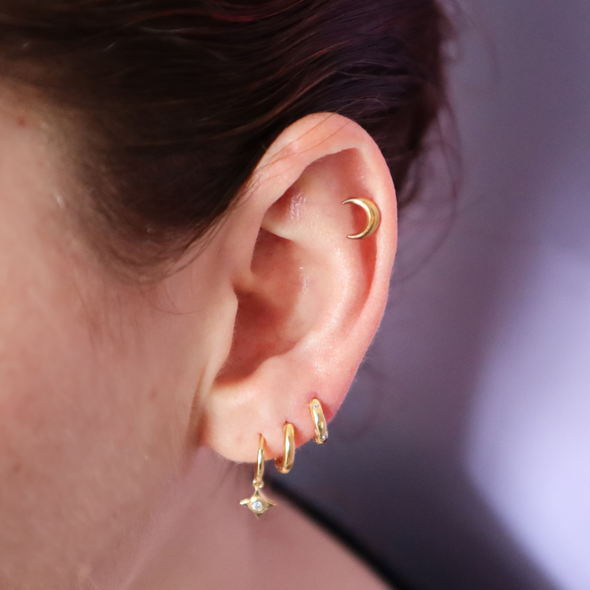 A close-up of an ear showcases the Papio Press Crescent Moon Labret Earring on the upper cartilage, paired with three small gold hoops—one featuring a tiny star charm—adorning the lower lobe.