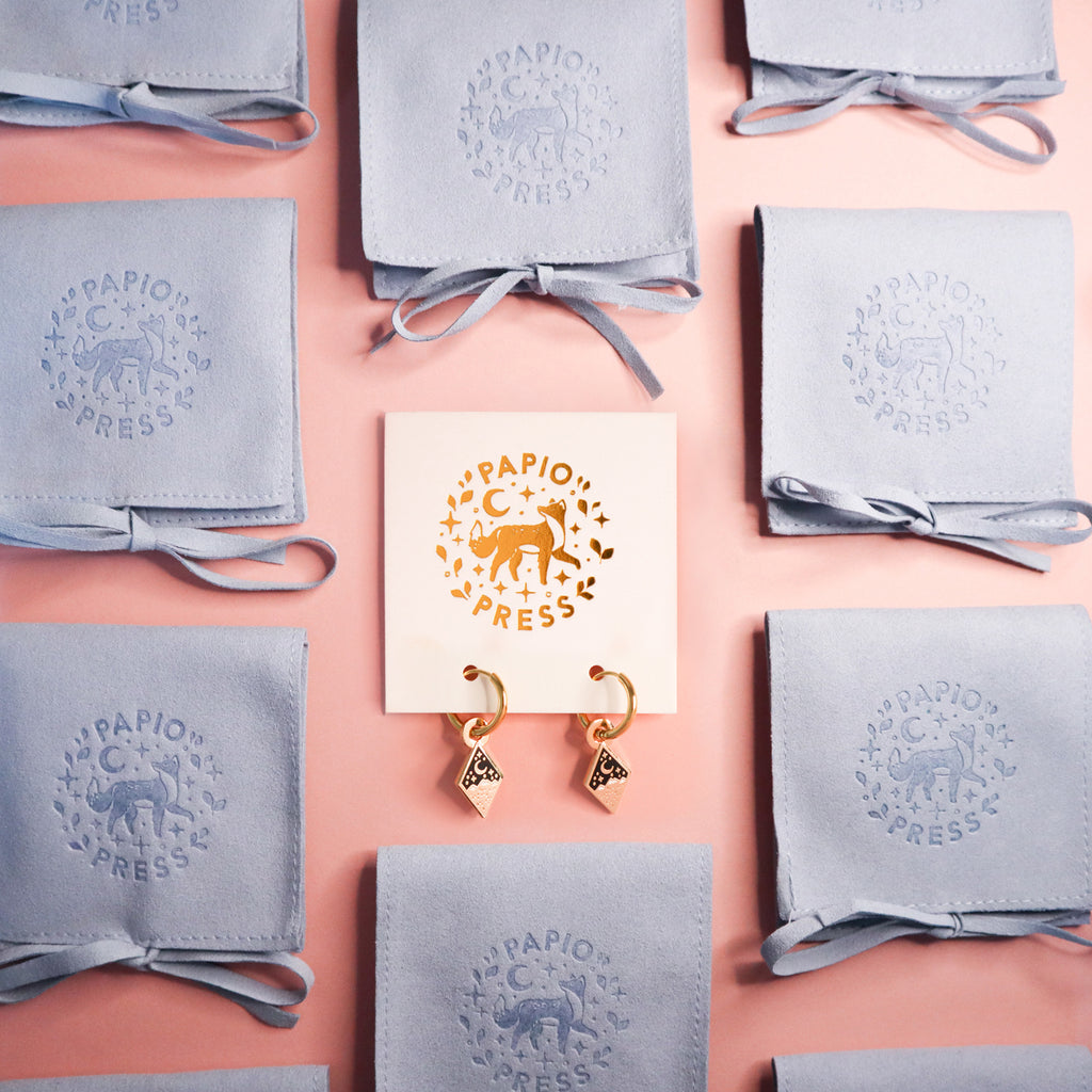 A photograph of a pair of gold mountain earrings on card backing board. They are surrounded by blue fabric Papio Press pouches on a pink background.