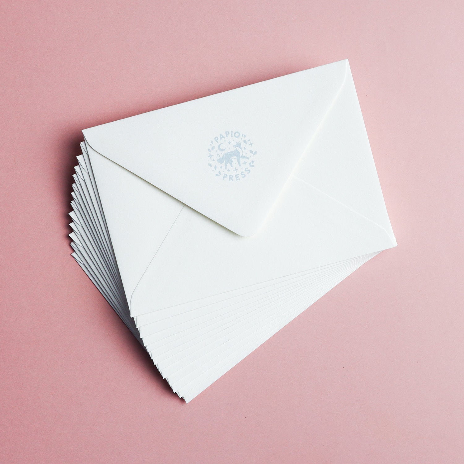 A stack of white envelopes with blue logos from Papio Press, arranged on a pink surface—perfect for sending the Thinking of You Owl Greetings Card.